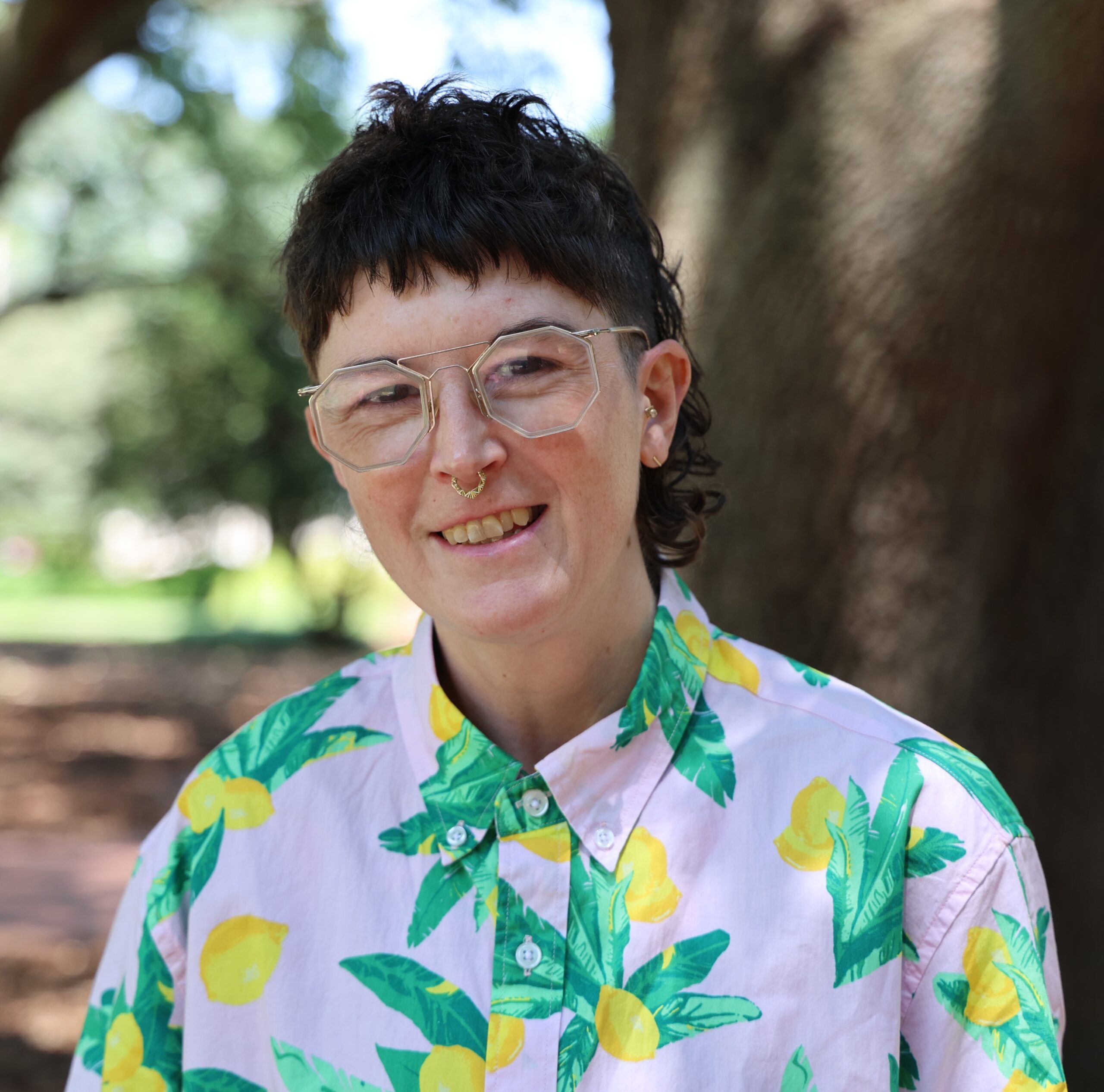 A person with short, textured hair and glasses stands outdoors, smiling. They are wearing a colorful shirt with a lemon and leaf pattern. The background features trees and greenery, suggesting a vibrant park setting.