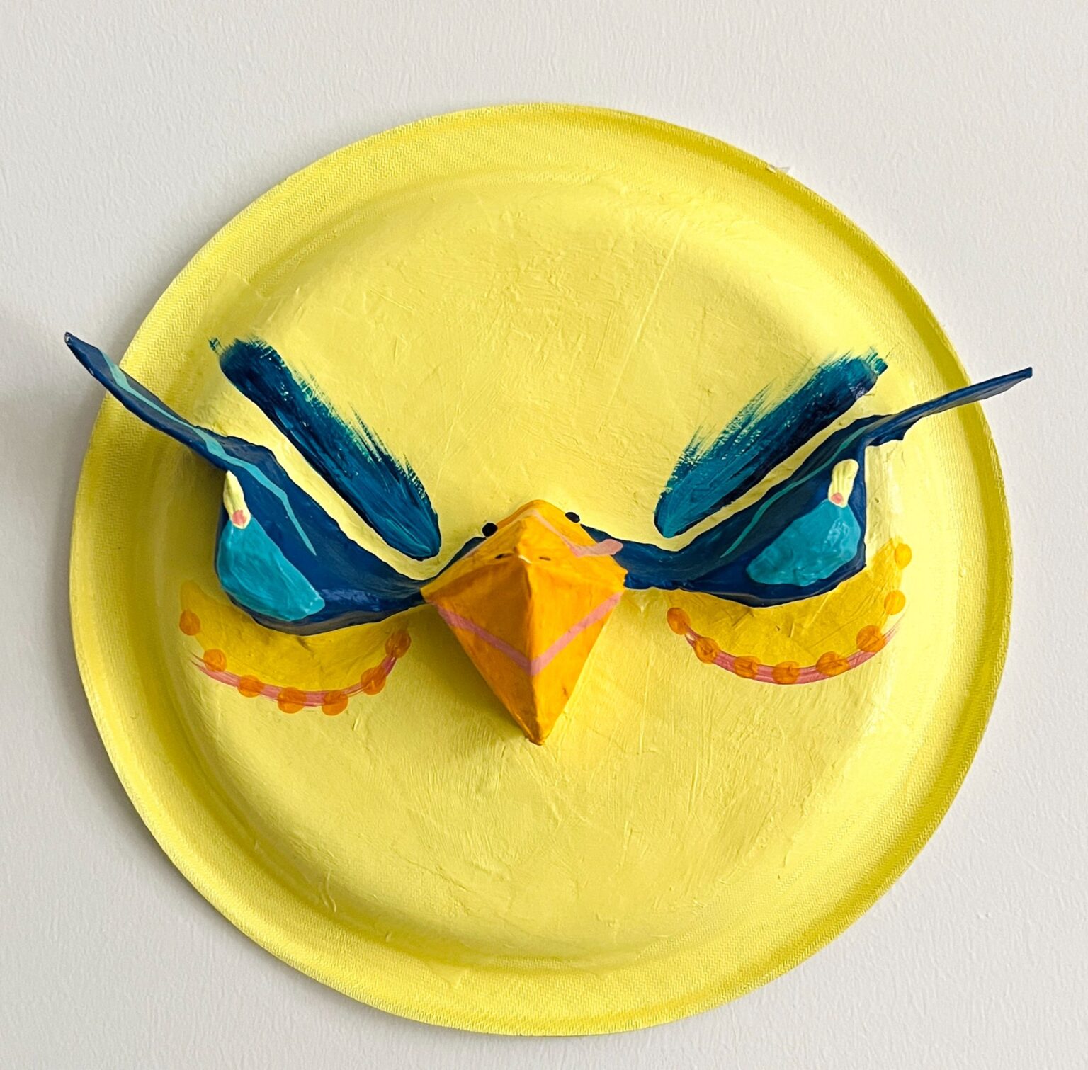 A brightly painted mask featuring a stylized bird face. The mask is painted yellow with a three-dimensional orange beak at the center, flanked by blue wings extending outward. Additional orange details suggest decorative plumage or markings.