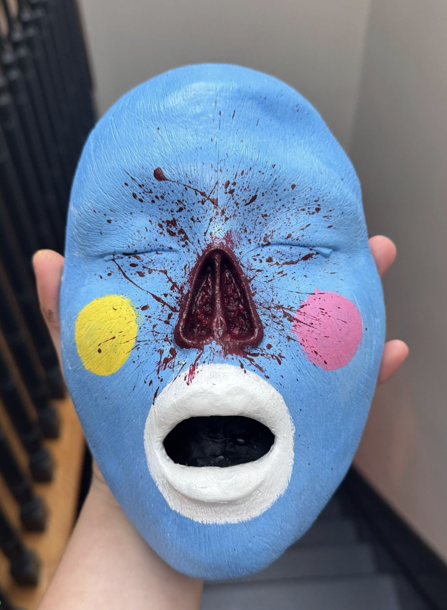A hand holds a stylized blue mask with closed eyes and an open mouth. The mask features a red splatter design at the center, with two colored circles—yellow on the left and pink on the right—on the cheeks. The nose is deep brown, contrasting with the mask's bright colors, and there appears to be a black interior visible through the mouth. The background consists of a staircase railing.