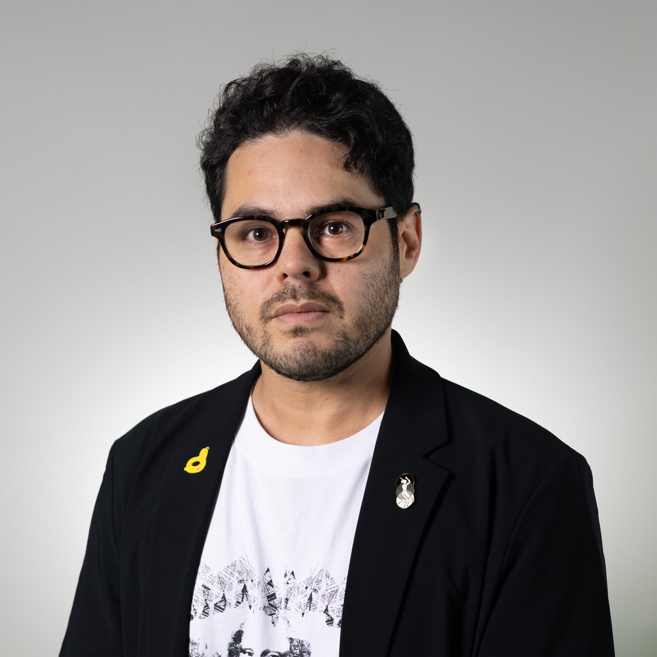 A person with short, curly hair and glasses is standing against a light gray background. They are wearing a black blazer over a white T-shirt featuring a graphic design. The blazer has two pins attached: a yellow one in the shape of a musical note and another silver pin. They appear serious and makes direct eye contact with the viewer.