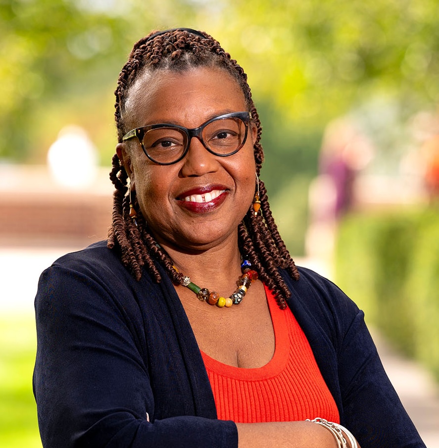 A person smiles at the camera, wearing glasses and a dark blue cardigan over a red top. They have braided hair adorned with colorful beads and a necklace made of various beads. The background features soft greenery and blurred figures, suggesting an outdoor setting.