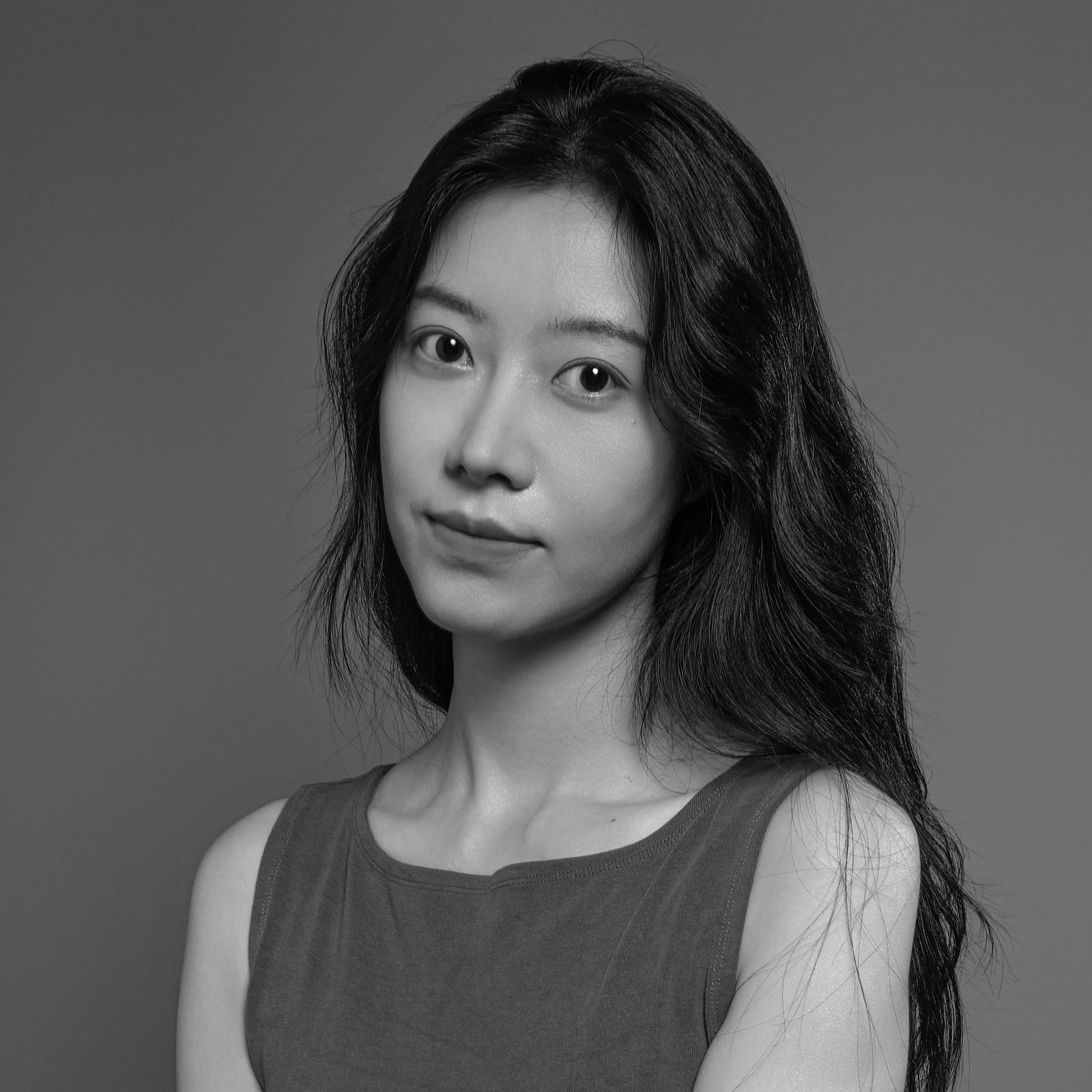 A young person with long, wavy hair is facing the camera, with a slight smile. They are wearing a sleeveless, dark top, and the background is a solid gray. The image is in black and white.