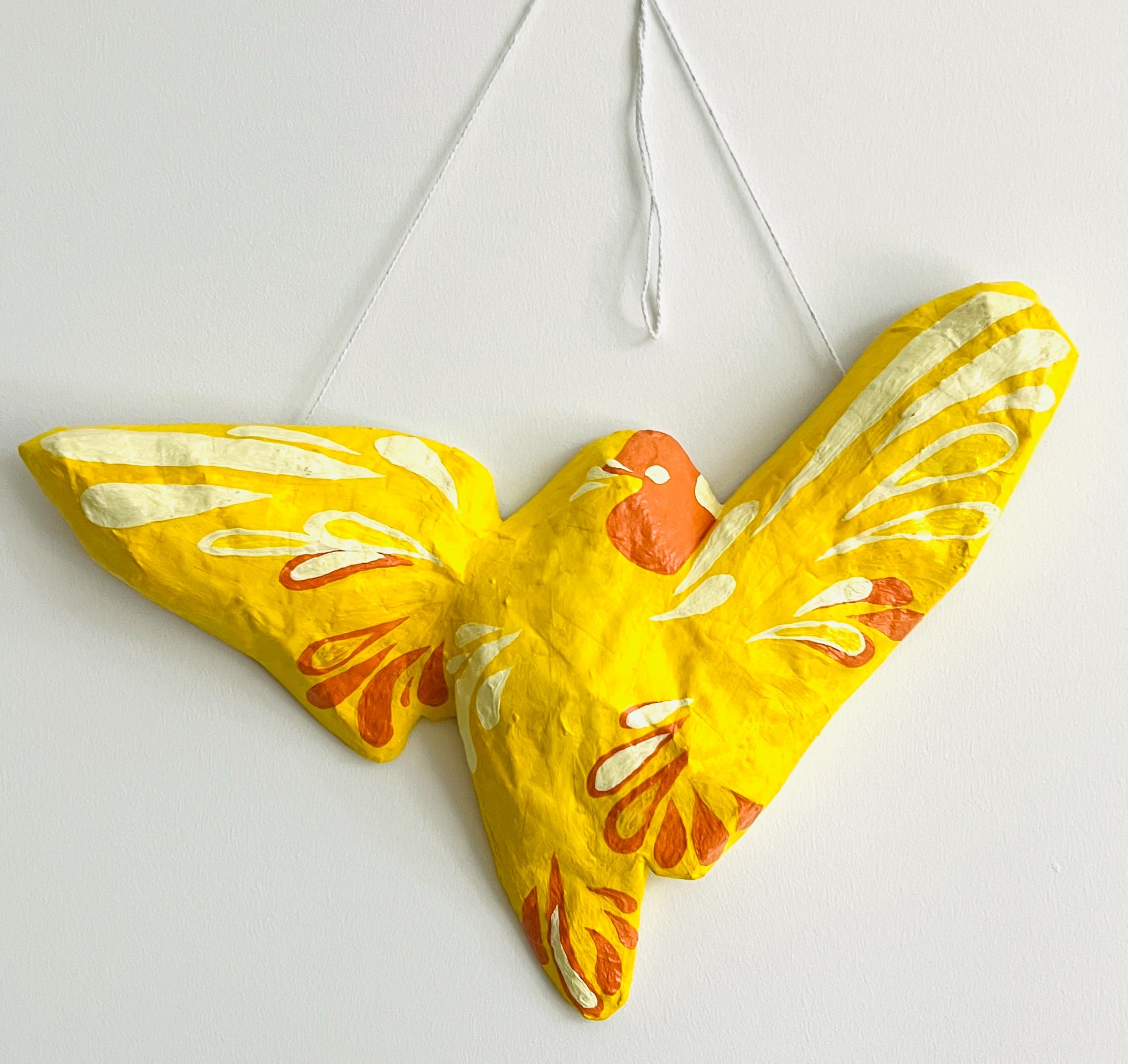 A brightly painted bird sculpture with outstretched wings, suspended by a white string against a white wall. The bird is primarily yellow with red and cream decorative markings, stylized to resemble feathers.