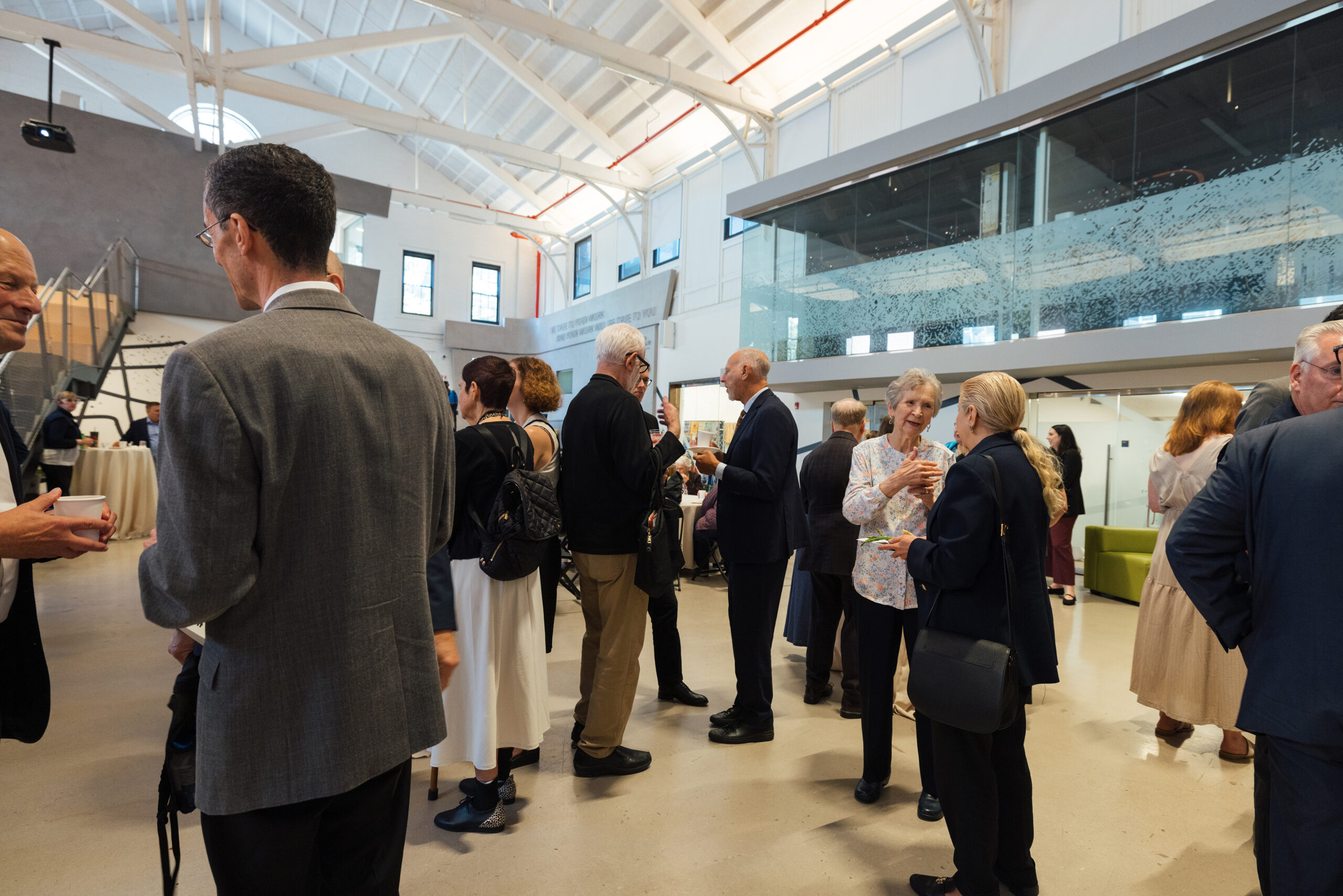 A group of people are mingling in a modern indoor space with high ceilings and large windows. Some individuals are engaged in conversations, while others hold drinks or food. The setting features various seating areas, and a few tables are visibly set up in the background. The atmosphere appears social and inviting.