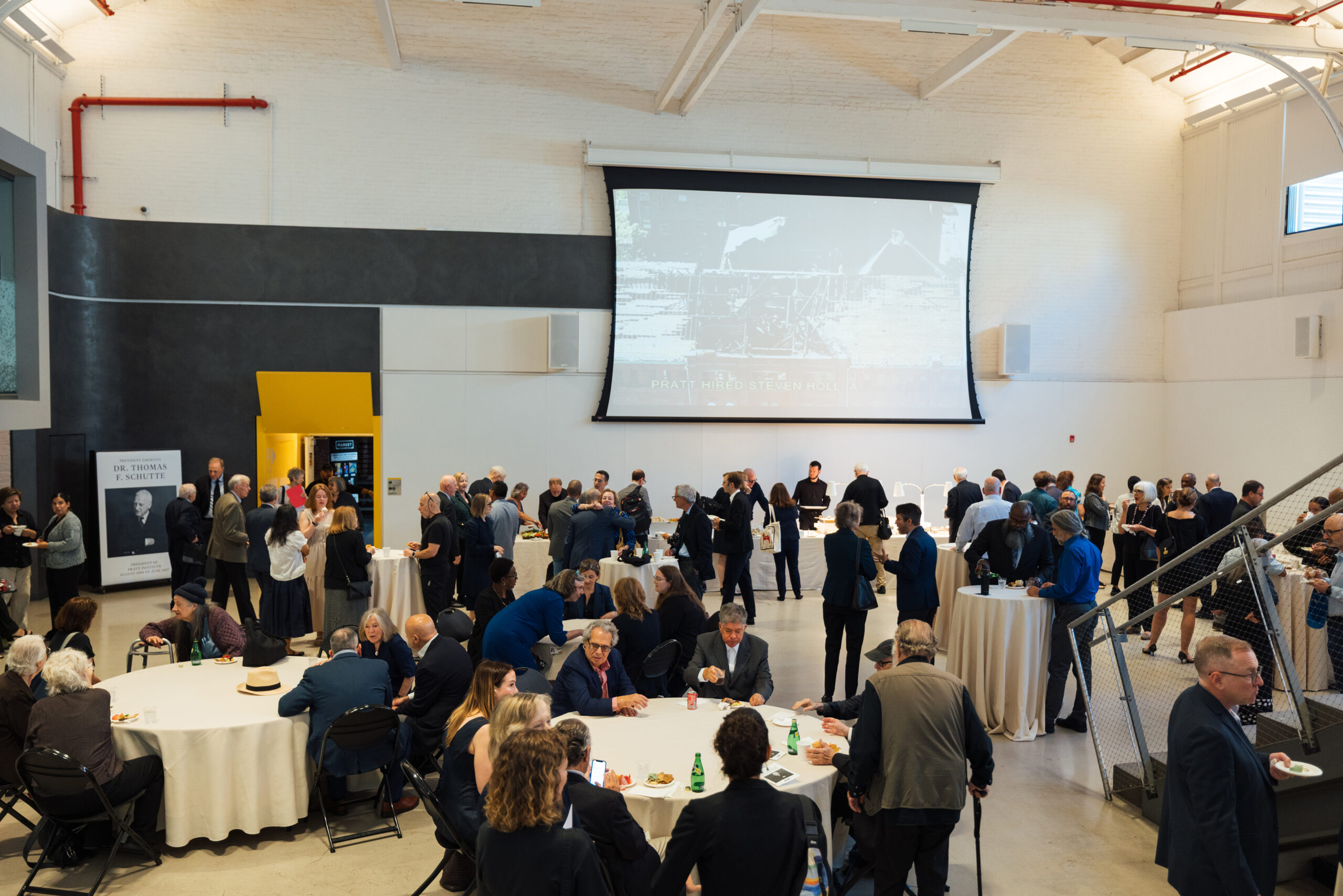 A bustling event space with guests mingling and engaging in conversation. Several tables are set up, some with food and beverages, while a large screen displays images. Attendees are dressed in formal and semi-formal attire, with a mix of genders and ages. In the background, a poster featuring an individual named Dr. Thomas F. Schutte is visible, along with a yellow doorway leading to another area. The atmosphere appears lively and social.