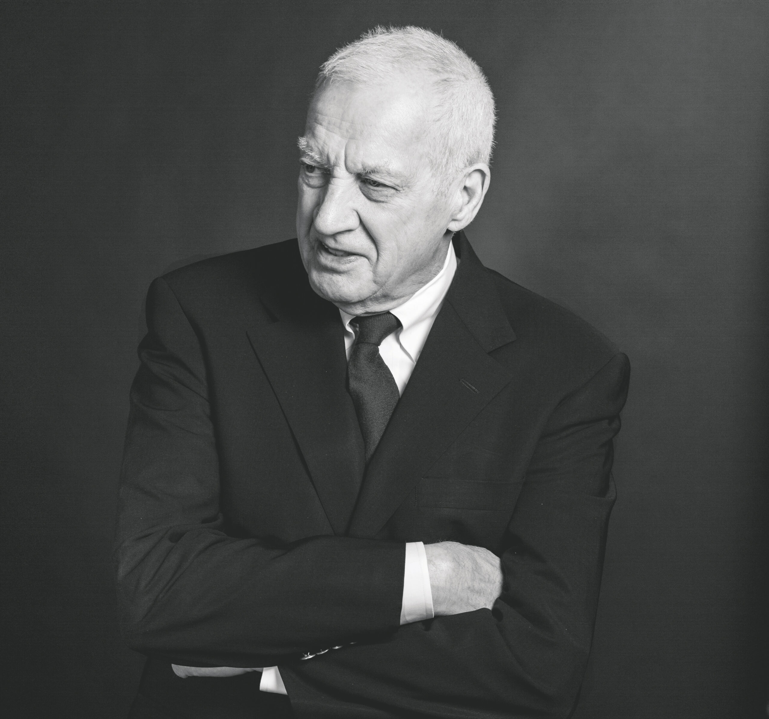 A man with gray hair is wearing a dark suit and tie, with his arms crossed. He has a serious expression and is looking slightly to the side. The background is a solid dark color, creating a formal atmosphere. The image is in black and white.
