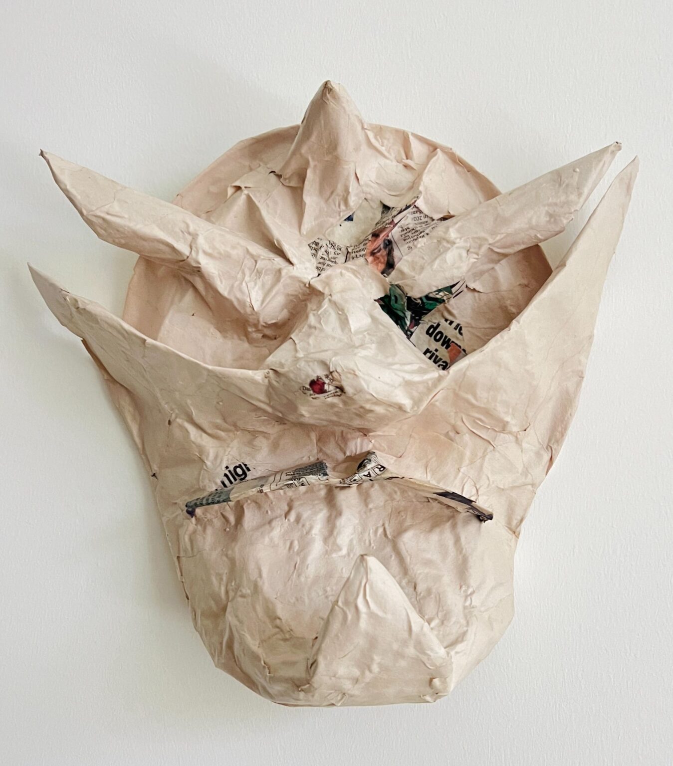 A papier-mâché mask made from layers of taped and crumpled paper, with pointed protrusions resembling horns or spikes. Areas of printed newspaper are visible through the beige surface, giving the mask a rough, unfinished texture.