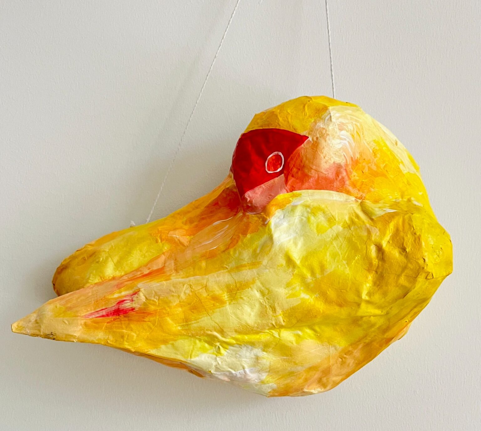 A papier-mâché bird sculpture painted in warm tones of yellow, orange, and red. The figure is shown in side profile, with a large rounded body, pointed beak, and a red patch around the eye. A string is attached for hanging.