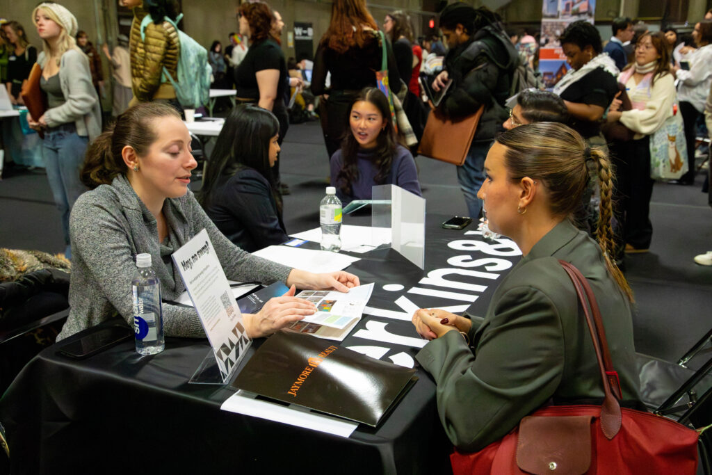 A group of people is gathered at a career fair or networking event in a large indoor space. At a table in the foreground, two women are engaged in a conversation; one is discussing materials while the other listens. Various attendees and booths are visible in the background, showcasing a bustling atmosphere. A water bottle and printed materials are on the table.