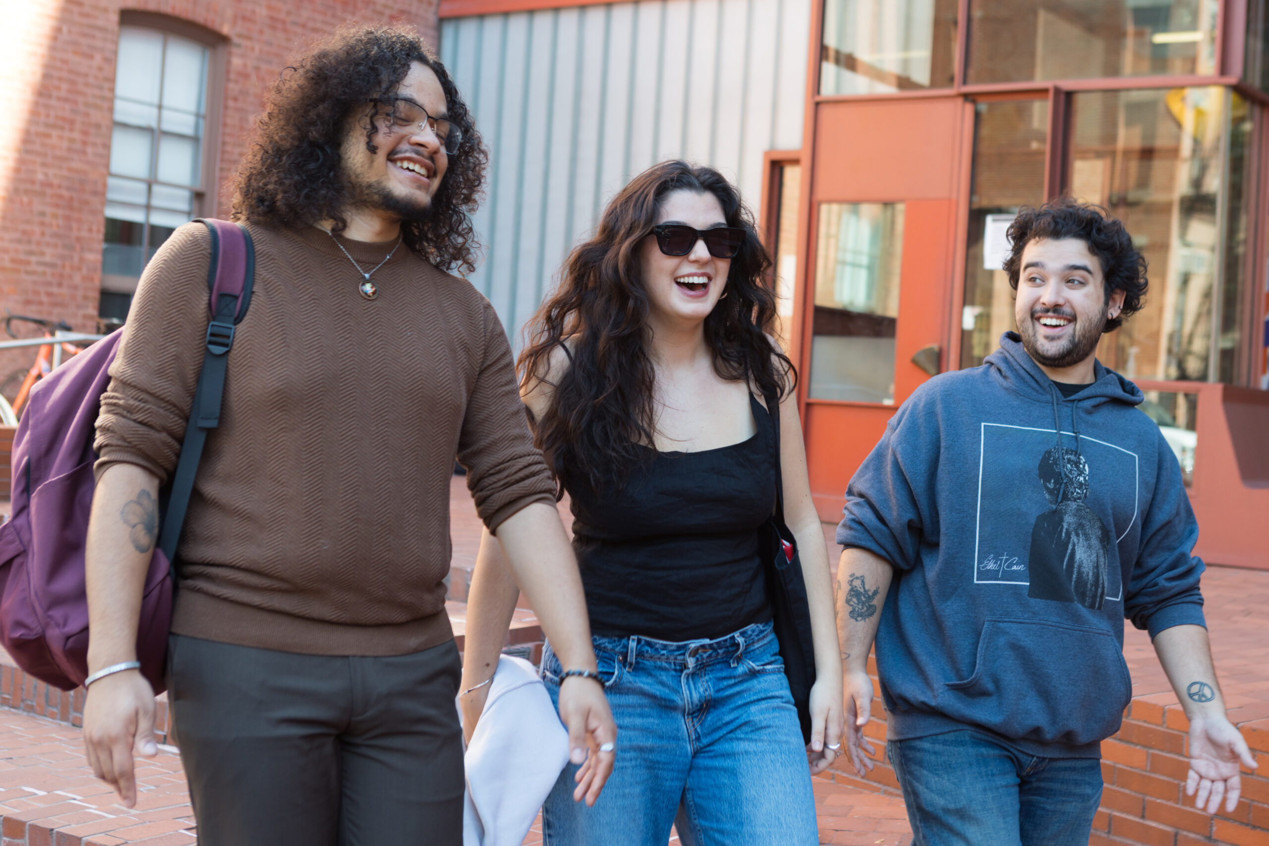 Three young adults are walking together and laughing outside. The person on the left has curly hair and a brown sweater with a backpack. The woman in the center has long, wavy hair and is wearing sunglasses and a black top, while the person on the right is in a blue hoodie and has short curly hair. They are in a brick setting with a glimpse of a building behind them.