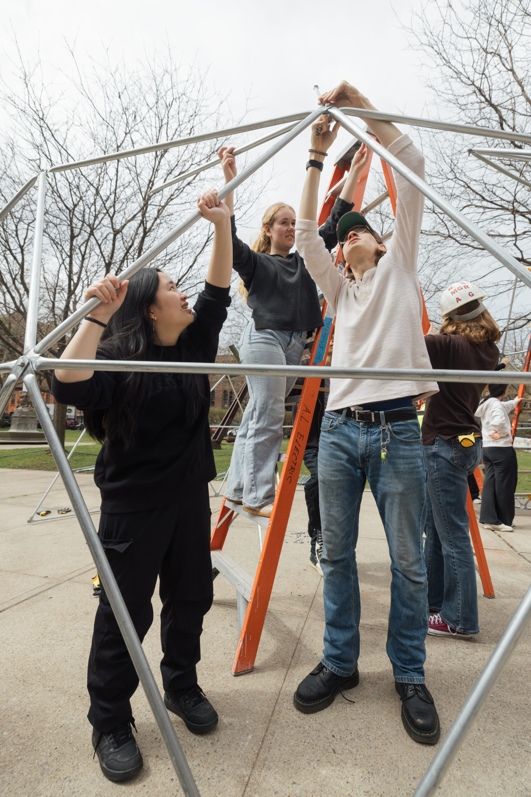 A group of people are constructing a geometric structure using metal rods. One individual on a ladder is holding a rod while others are positioned around the base, helping to connect the pieces. Two people in the foreground are gripping the rods, while another person stands on the ladder in the background. The scene takes place outdoors amid bare trees and a concrete surface. The sky is cloudy, indicating overcast weather.