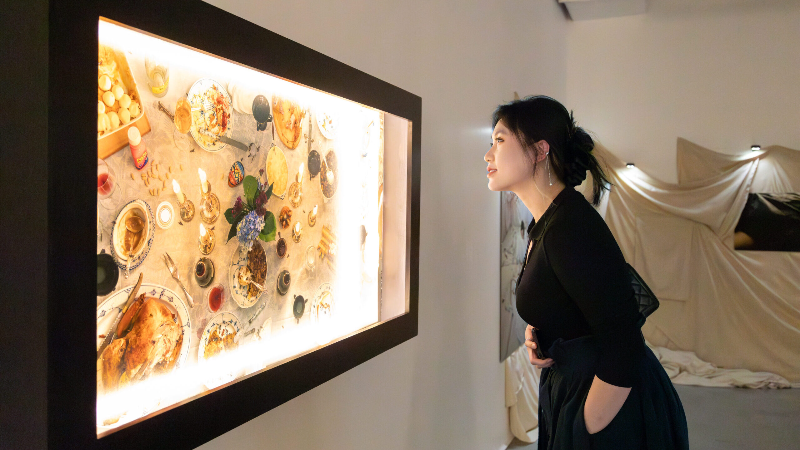 A woman gazes at a brightly lit display, creating a festive atmosphere. She is dressed in a black top and skirt, standing in a minimalist gallery space with soft lighting and fabric draping in the background.