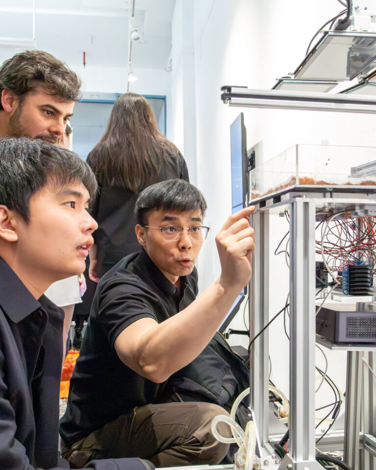 Three individuals are gathered around a scientific apparatus with a transparent container at its center. One person, wearing glasses and a black shirt, is pointing and explaining something, while the other two are listening intently. In the background, there are electronic devices and wires, indicating a technical or research setting. The atmosphere appears collaborative and focused.
