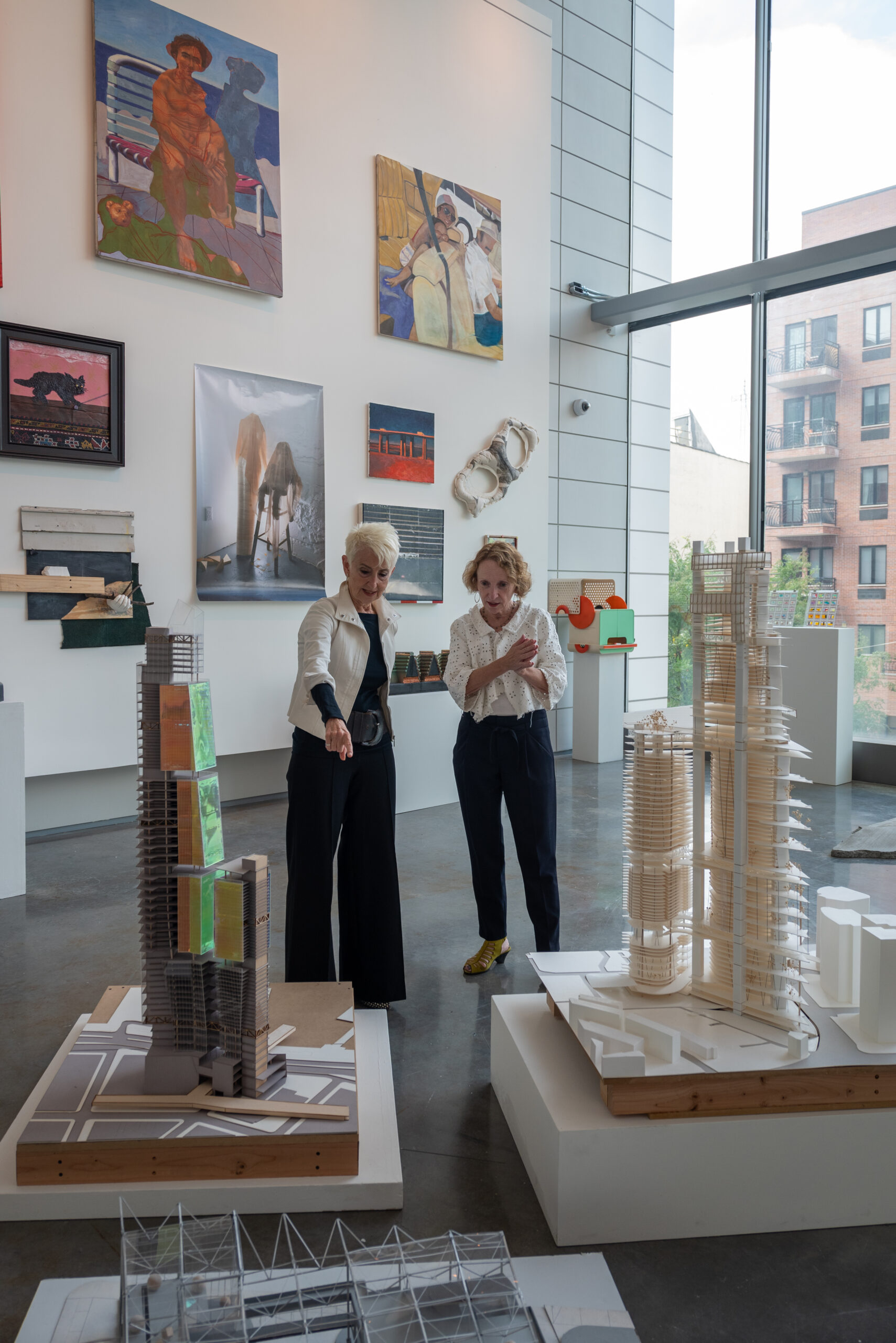 Two women stand in an art gallery, admiring architectural models displayed on pedestals. One woman gestures toward a tall model with colorful sections, while the other claps her hands in excitement. Various artworks adorn the gallery walls, including paintings and mixed media pieces. Large windows provide natural light and a view of buildings outside. The scene conveys a sense of engagement and creativity in a modern space dedicated to art and architecture.