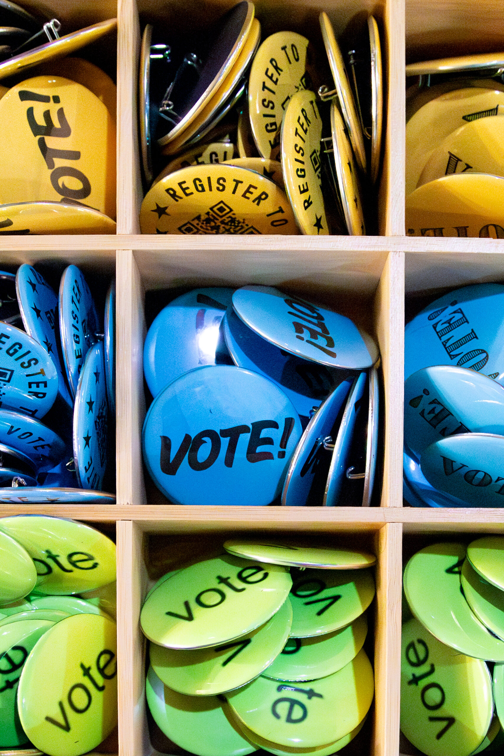 Close-up of colorful buttons organized in a wooden display grid. The buttons are yellow, blue, and green, printed with words like “VOTE!” and “REGISTER TO VOTE,” promoting civic engagement and voter registration.