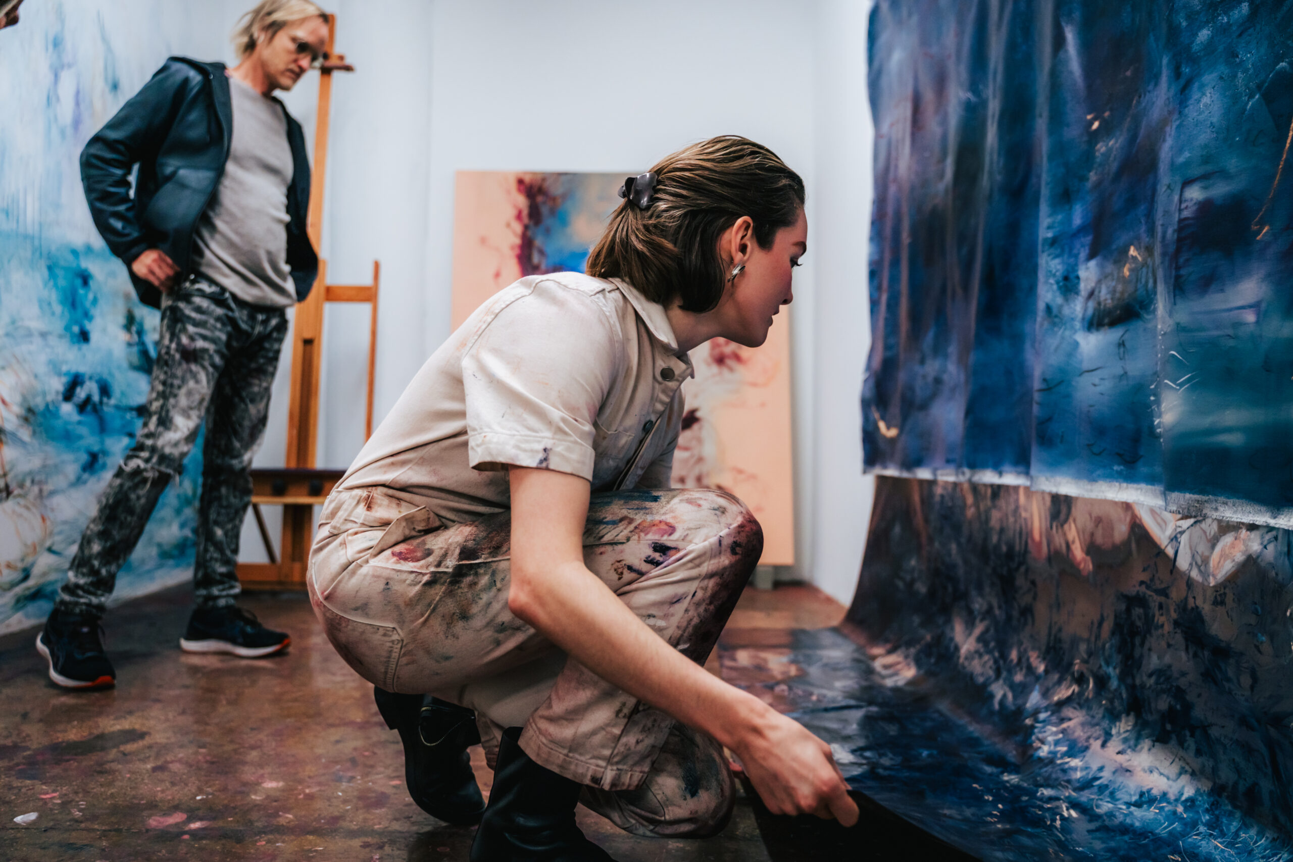 A person with short hair, dressed in a light-colored, paint-splattered jumpsuit and black boots, is crouched down, examining a large piece of blue-toned artwork. Another individual, wearing a dark jacket and patterned pants, stands in the background, observing. The studio has various artworks displayed, and the setting has a creative, artistic atmosphere.