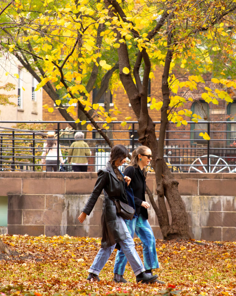 Two women are walking side by side through a park covered in fallen autumn leaves. One woman, wearing a black leather jacket, has dark hair and jeans, while the other, dressed in a black top and blue jeans, has her hair tied back. In the background, trees with yellow leaves can be seen, along with people walking and sitting on benches.