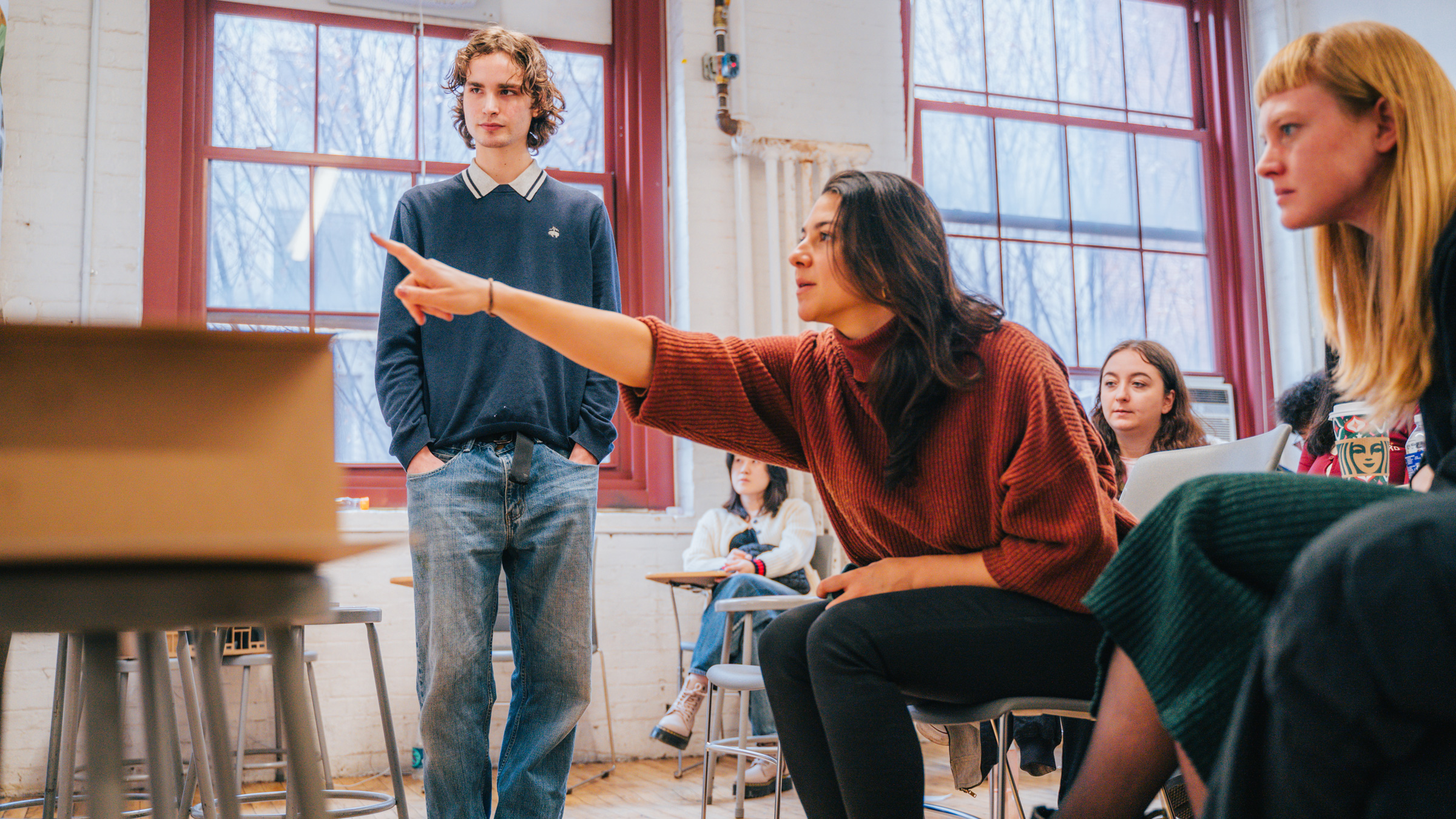 A group of students in a classroom setting participate in a discussion. A woman seated in front gestures expressively while talking, wearing a red sweater. A man stands nearby with his hands in his pockets, observing. Other students are seated in chairs, listening attentively, with some visible reactions. Large windows in the background allow natural light into the room.