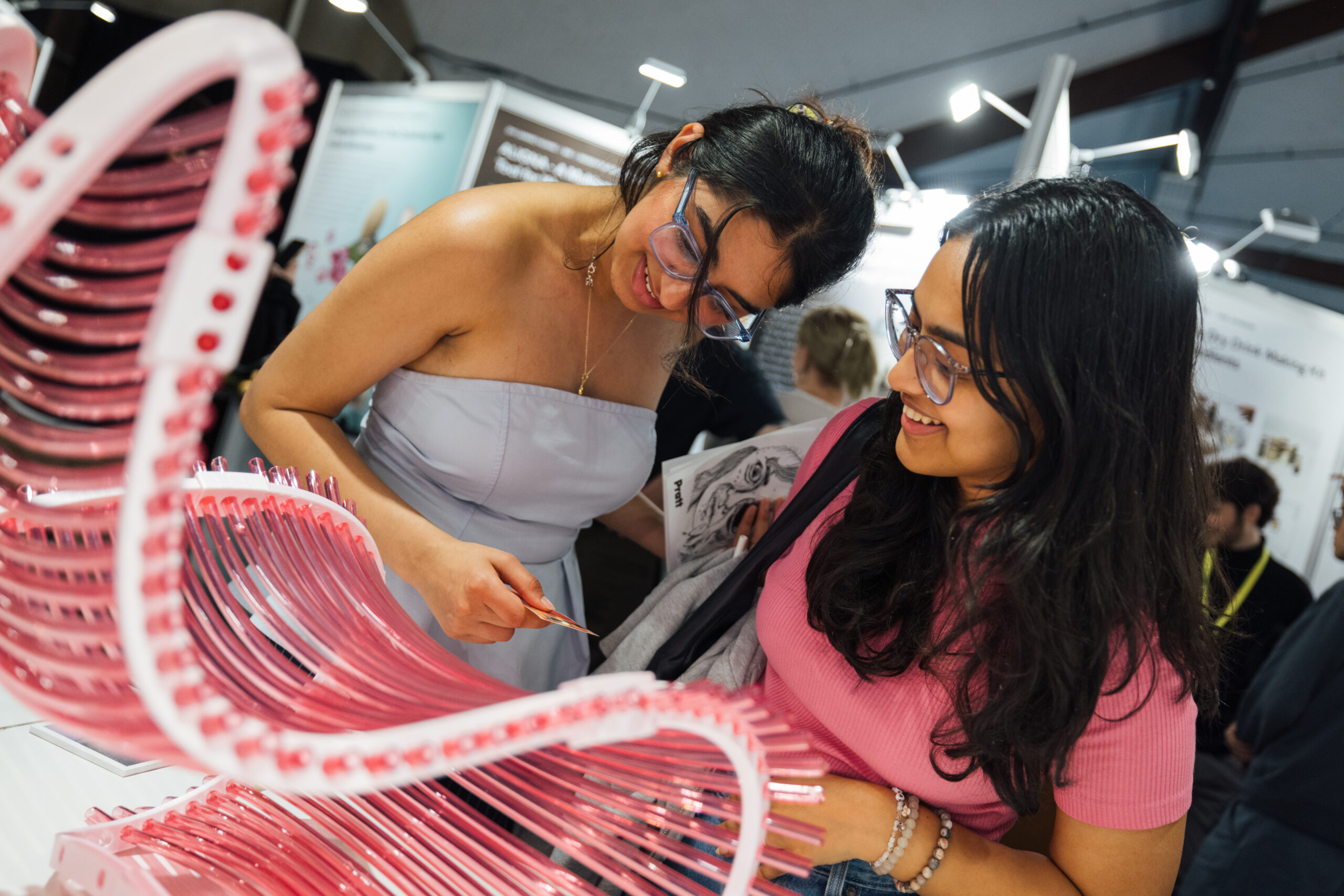 Two women are examining a pink, curved display with various clear sections, likely part of an exhibition. One woman, wearing a dress and glasses, points at the display while the other, in a pink top and glasses, smiles as she looks on. Background displays and other attendees can be seen blurred behind them.