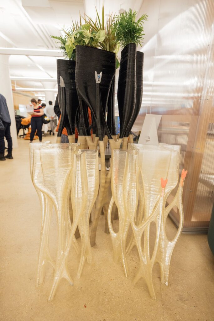 A collection of uniquely designed vases, some tall and black with leafy plants, and others shorter and translucent, feature a wavy, organic shape. The vases are arranged together in a workspace setting. People are visible in the background, engaged in various activities. The floor is light-colored concrete, and the workspace is well-lit with modern architectural elements.