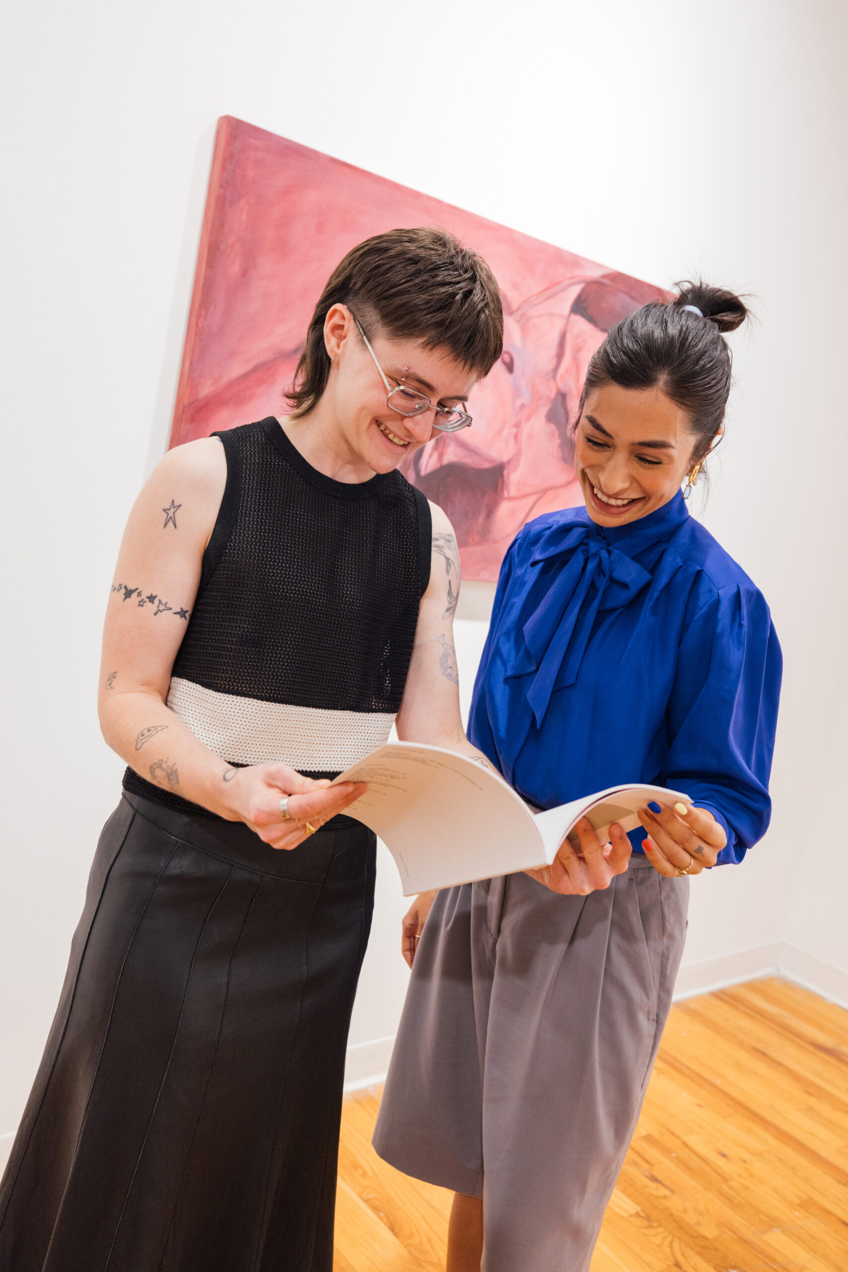 Two individuals are engaging in conversation while looking at a book in an art gallery. One person has short hair and glasses, wearing a black sleeveless top with a white panel and a long black skirt. The other has long, dark hair in a ponytail, dressed in a bright blue blouse with a bow tie and light gray pants. A large pink artwork is visible in the background.