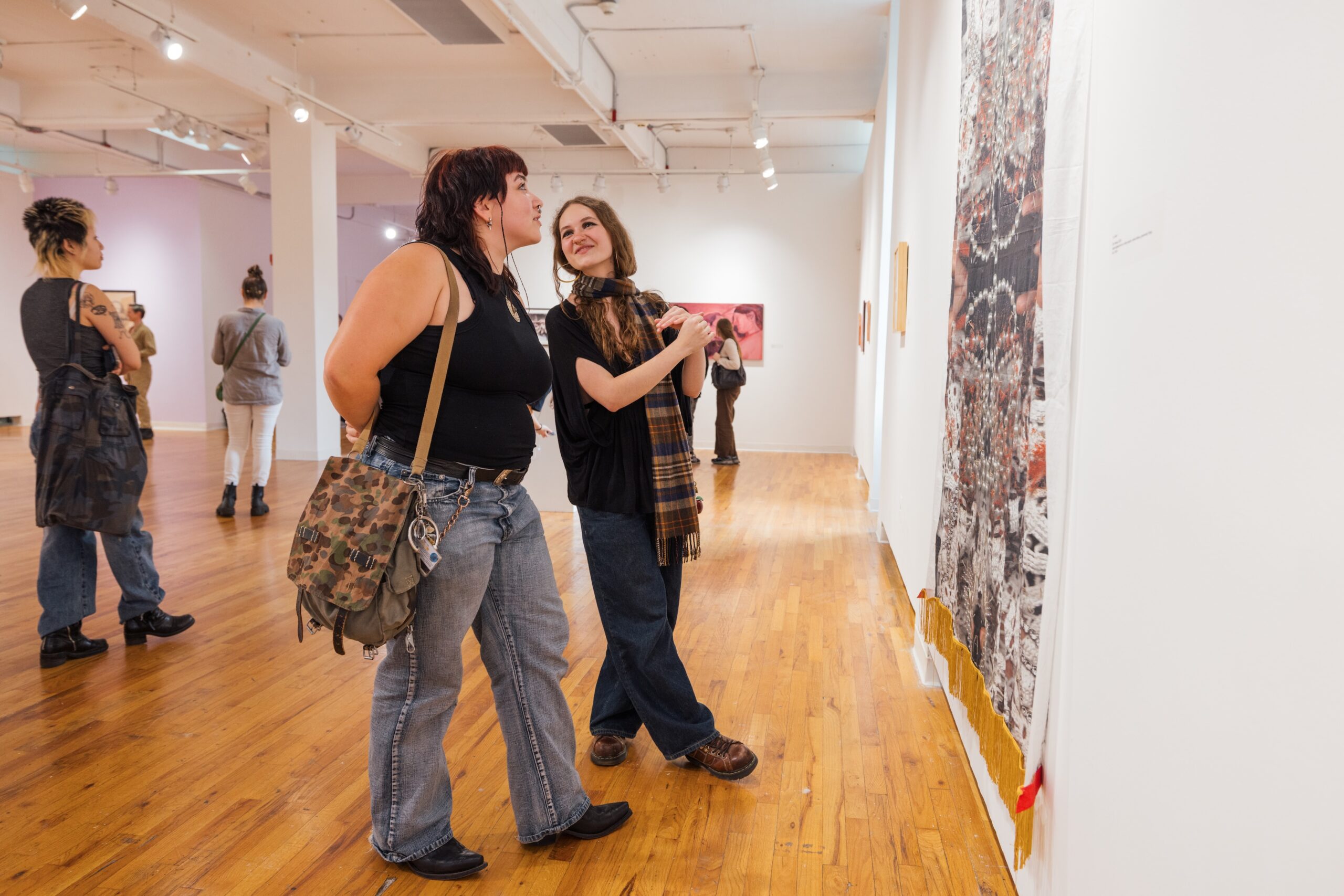 Two people are engaged in conversation while looking at an artwork in an art gallery. They appear to be smiling and enjoying the moment. The background includes other visitors exploring the gallery, with various art pieces displayed on white walls. The flooring is polished wood, enhancing the gallery's welcoming atmosphere.