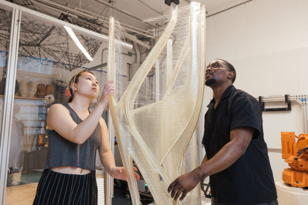 Two individuals are working together in a studio space. One person, a woman with short hair, holds a translucent, sculptural piece while looking upward, concentrating on its position. The other person, a man wearing glasses, is also looking up and is positioned next to her, assisting. The background features various tools and artistic items, suggesting a creative environment.