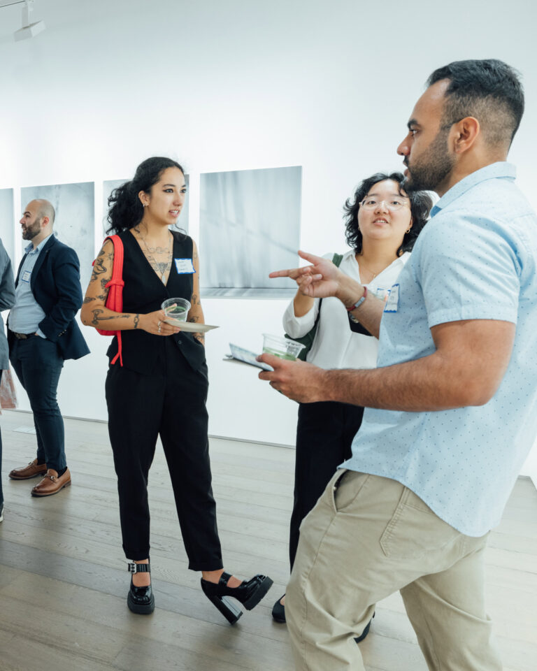 A group of four people engages in conversation at an art gallery. One woman with tattoos and wearing a black outfit stands with her arms crossed. Another woman, dressed in white, listens attentively while gesturing. A man in a light blue shirt holds papers and appears to be speaking. In the background, other attendees mingle by several monochromatic artworks displayed on the wall. The setting features light wood flooring and bright, modern lighting.