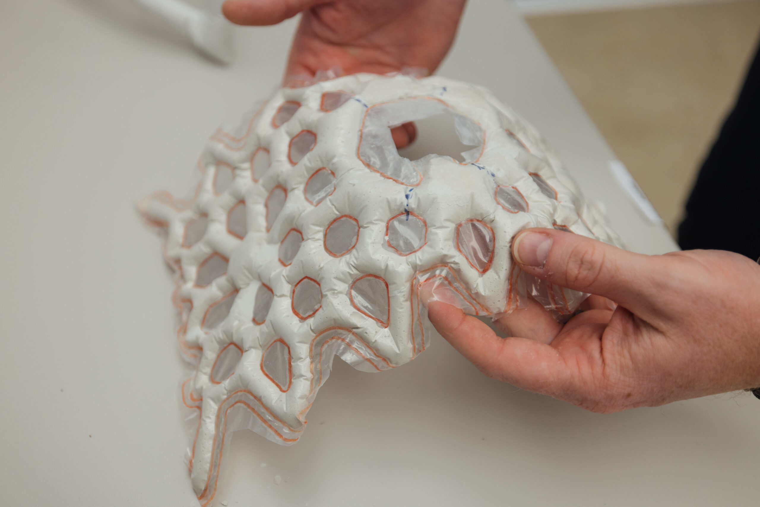 Close-up of a person holding a flexible, geometric architectural prototype made of white material with translucent hexagonal cutouts outlined in orange. The structure has a lightweight, inflated or molded texture and appears to be part of an experimental design or materials research project, photographed against a neutral background.