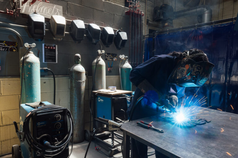A person wearing a protective welding helmet is working on a metal surface, using a welding tool that creates bright sparks and blue light. In the background, there are multiple gas cylinders and a welder machine. The workspace features various tools and safety equipment, with a blue curtain separating the welding area from other parts of the shop.