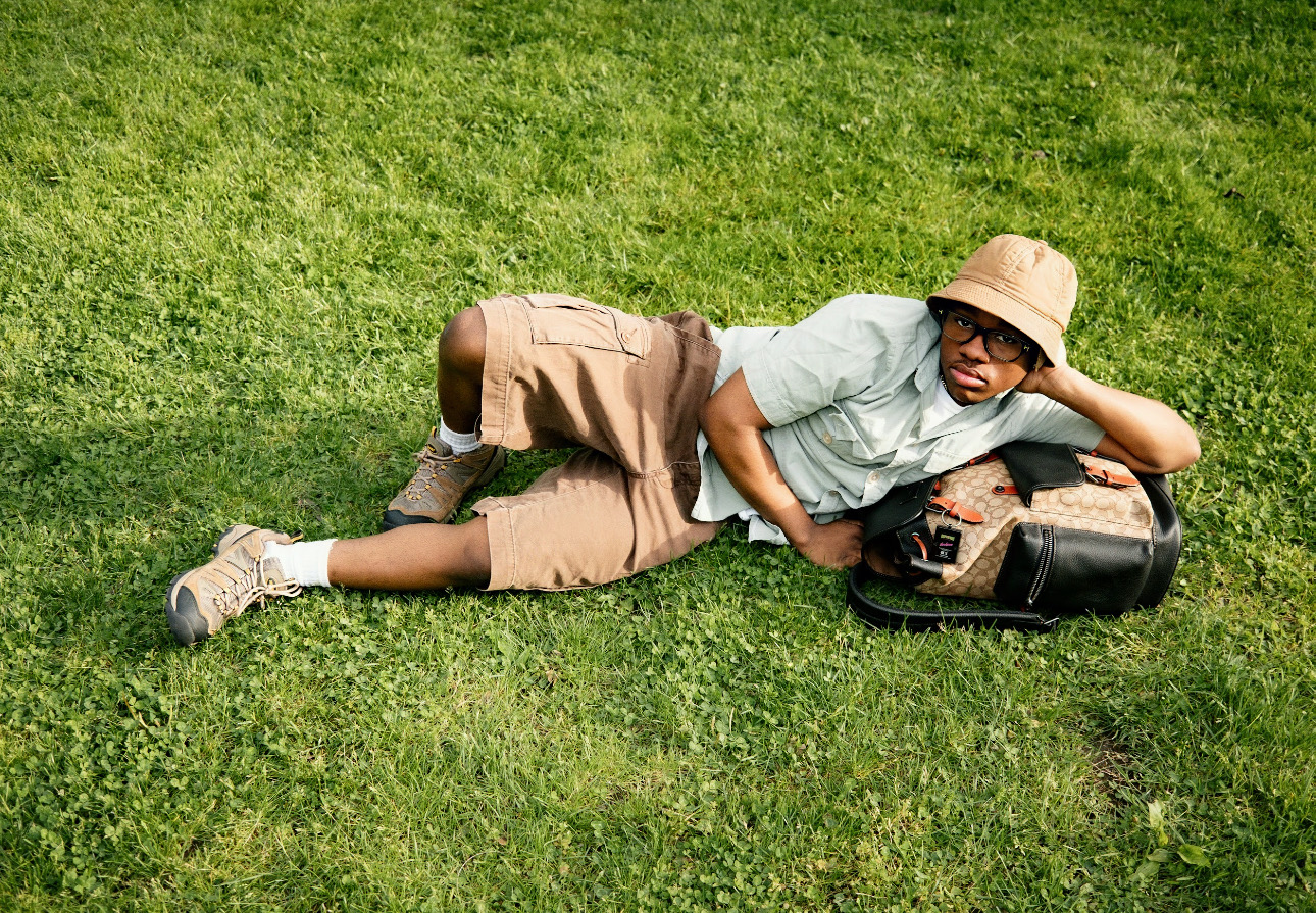A person is lying on green grass, resting on one arm with a casual expression. They are wearing a light green short-sleeve shirt, tan shorts, and hiking shoes. A light beige bucket hat sits on their head. Beside them is a large bag with a mix of brown and black colors. The setting is bright and outdoors, emphasizing a relaxed vibe.