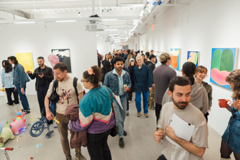 A crowded art gallery scene with diverse attendees interacting, looking at artworks on white walls. Some people are holding drinks or taking notes, while others are chatting. Colorful abstract art pieces are displayed prominently. The gallery is bright with a modern aesthetic, featuring a mix of casual and stylish clothing among the visitors. Discarded objects can be seen on the floor, indicating an interactive or lively atmosphere.