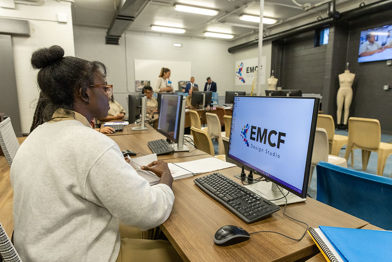 A student with glasses sits at a computer in a design studio, focused on the screen displaying the EMCF Design Studio logo. The classroom features multiple computers, chairs, and mannequins in the background. Two people are visible in the distance, engaging with each other. The environment is well-lit and organized, showcasing a learning space geared toward design education.