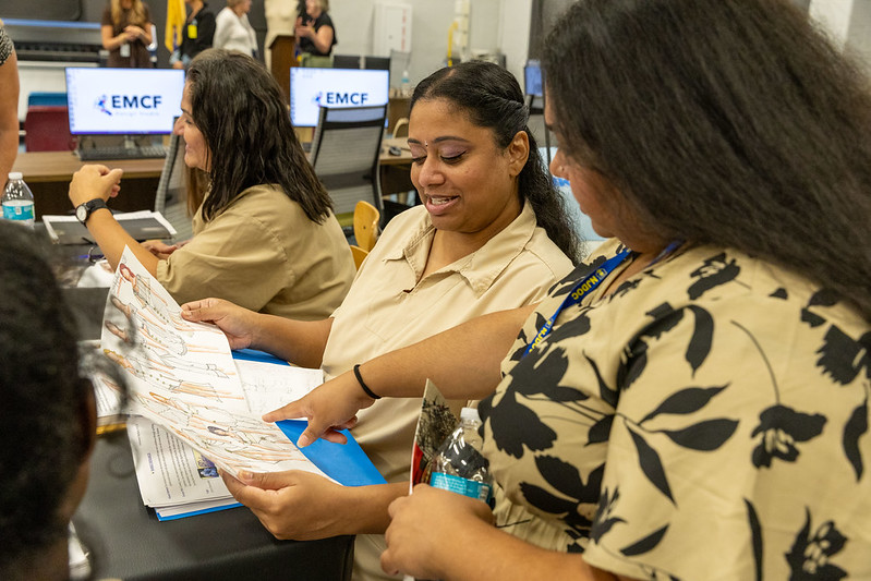 Two women are discussing a map or document while seated at a table. One woman is pointing to the document, while the other woman is smiling and looking at it. Both are wearing light-colored shirts, and there are additional people and computer screens visible in the background. The setting appears to be an office or training environment.