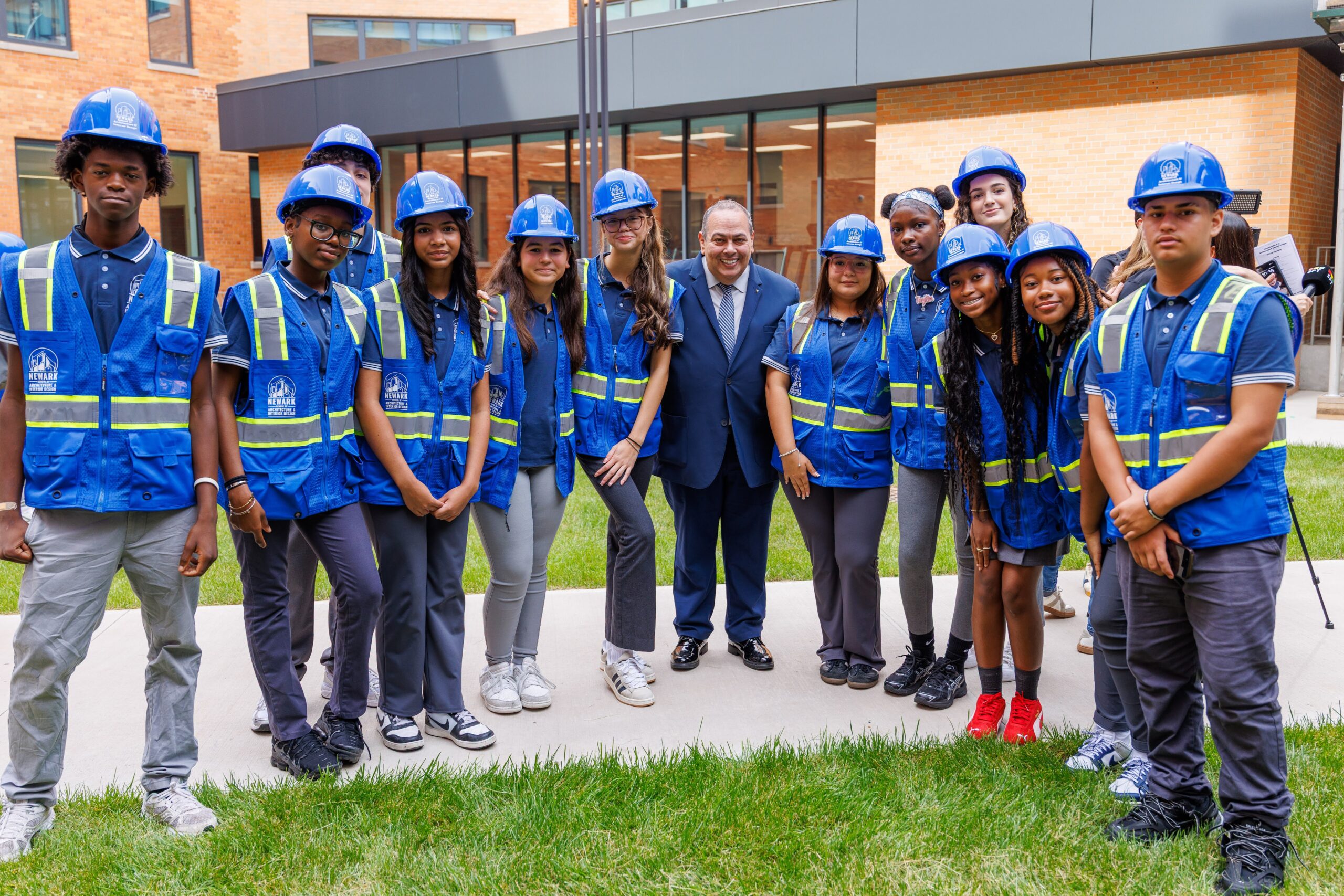 A group of twelve students wearing blue hard hats and safety vests stands outdoors, smiling. They are gathered around a man in a suit, who is also smiling. The backdrop features a modern building with large windows and green grass in the foreground. The students appear engaged and enthusiastic about the event.
