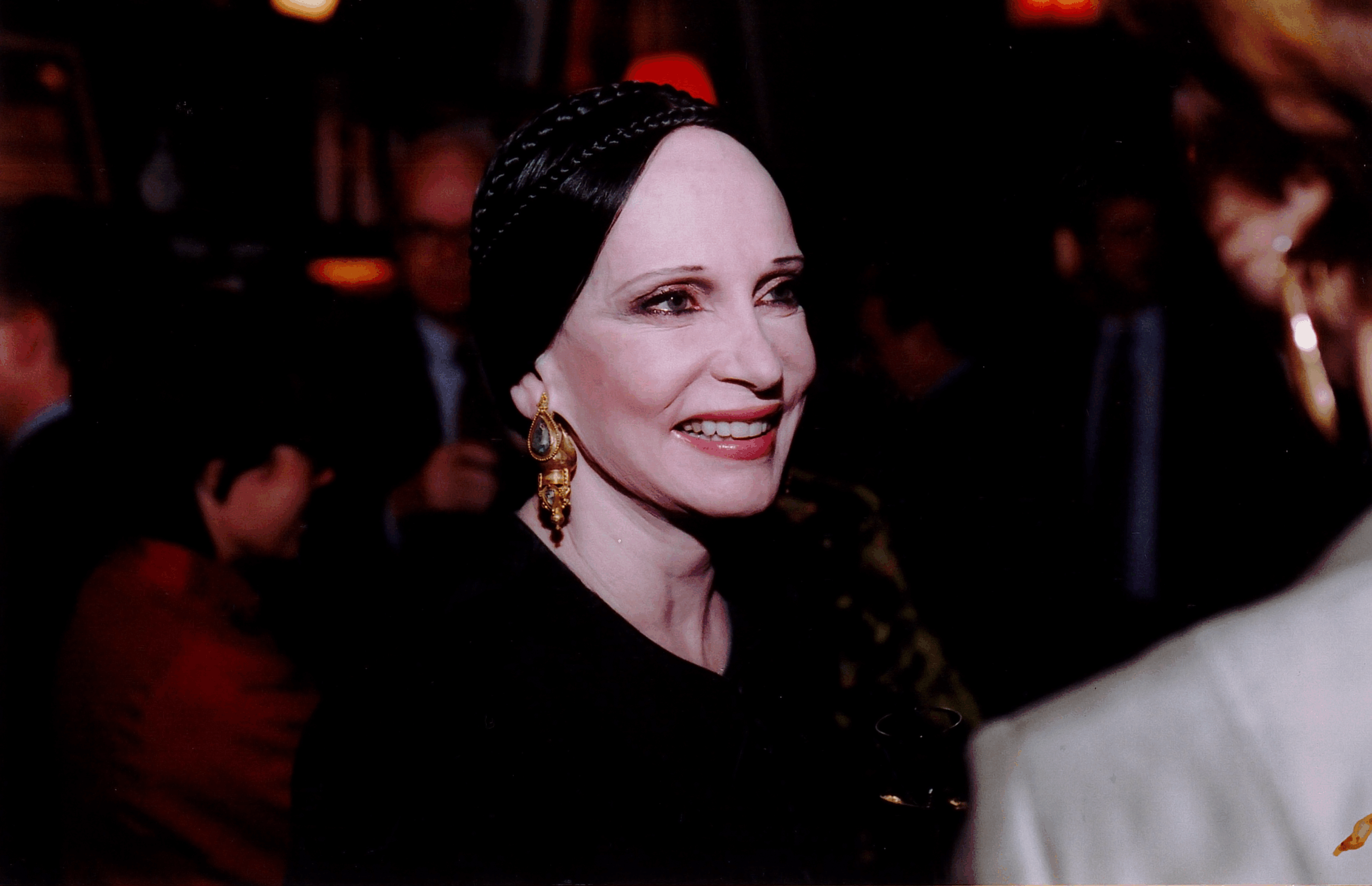 A woman with dark hair styled in a braid is smiling, wearing large golden earrings and a black outfit. She is engaged in conversation, and people are blurred in the background, suggesting a social setting or event. The lighting is dim, adding to the intimate atmosphere of the gathering.