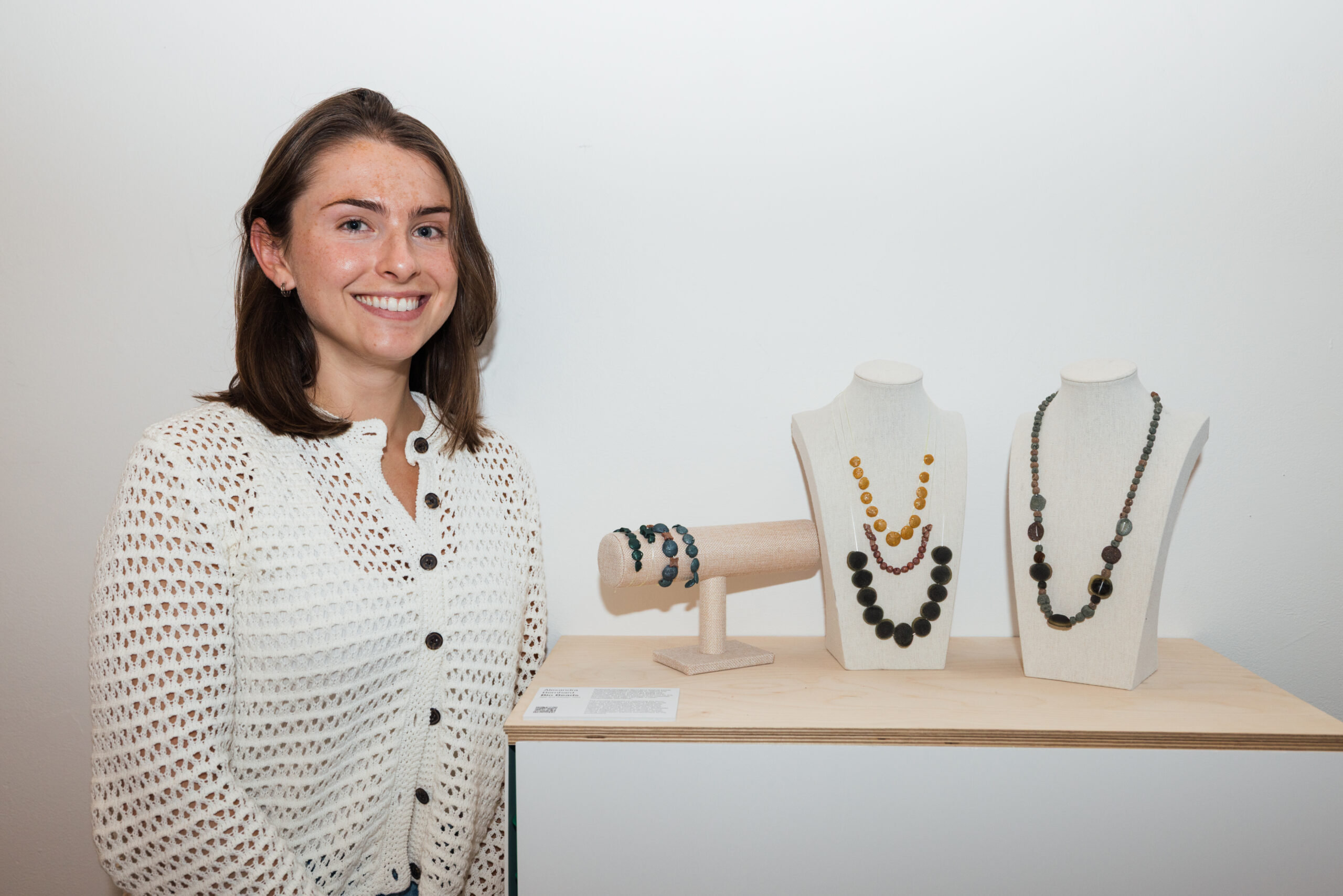 A smiling individual stands beside a display of artistic sculptures in an exhibition space. They wear a dark, intricately designed sleeveless top and light-colored trousers. The background features tall metal sculptures alongside smaller pieces mounted on stands, showcasing varied materials and textures. A wooden door is partially visible to the right.
