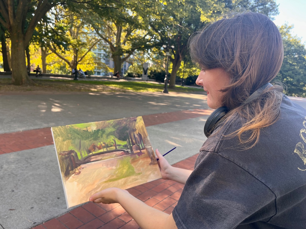 A person sits on the ground beneath a tree, surrounded by fallen leaves and grass. They are smiling and holding a painting or drawing on a clipboard. Art supplies are scattered nearby on a blanket, and the tree's thick trunk and roots are visible in the foreground. The scene conveys a peaceful outdoor art activity.