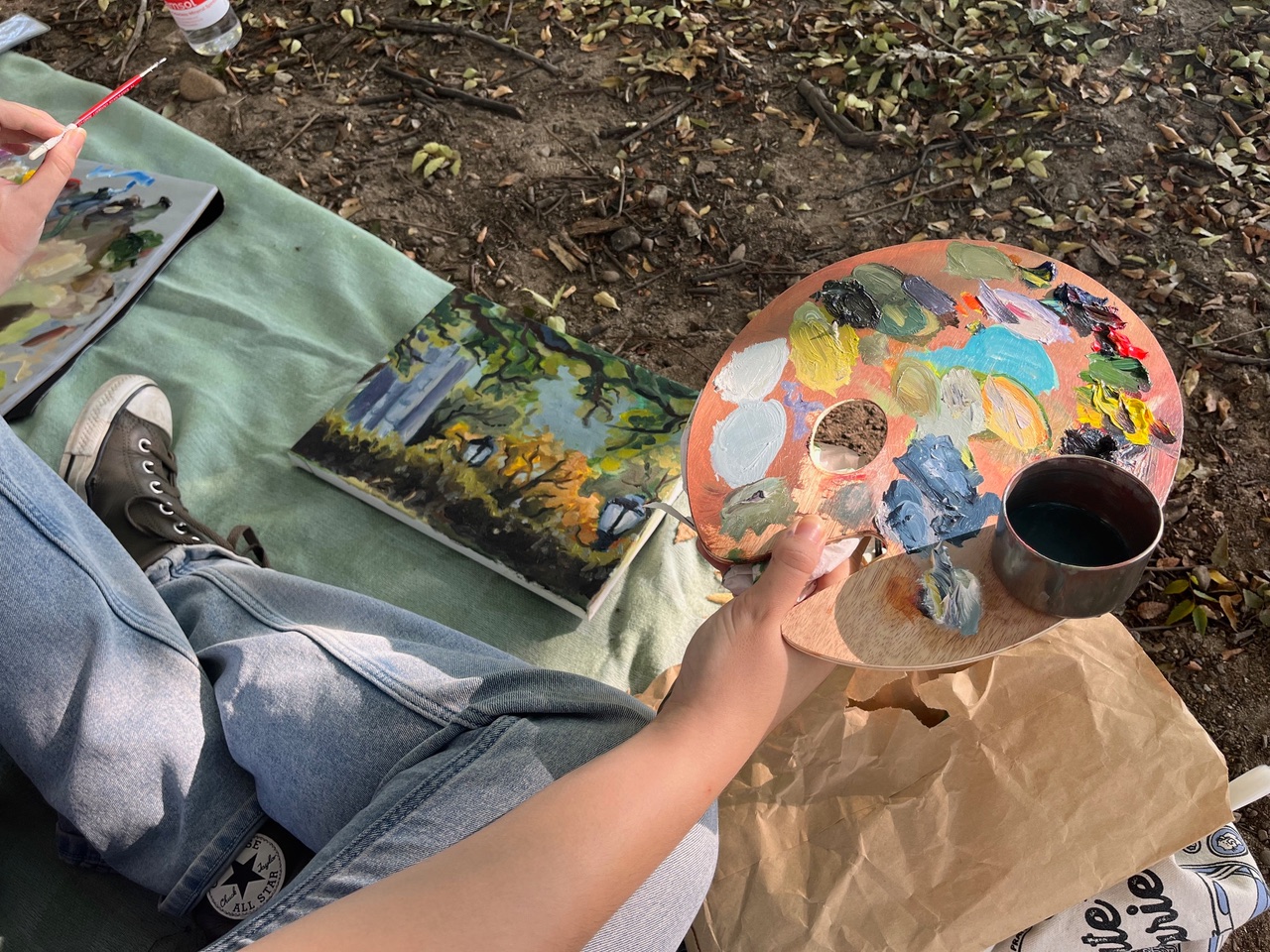 A person sitting on the ground under a large tree, smiling while holding a paintbrush and a canvas. They are surrounded by fallen leaves and have art supplies scattered nearby. The setting is a calm, natural area with greenery in the background.