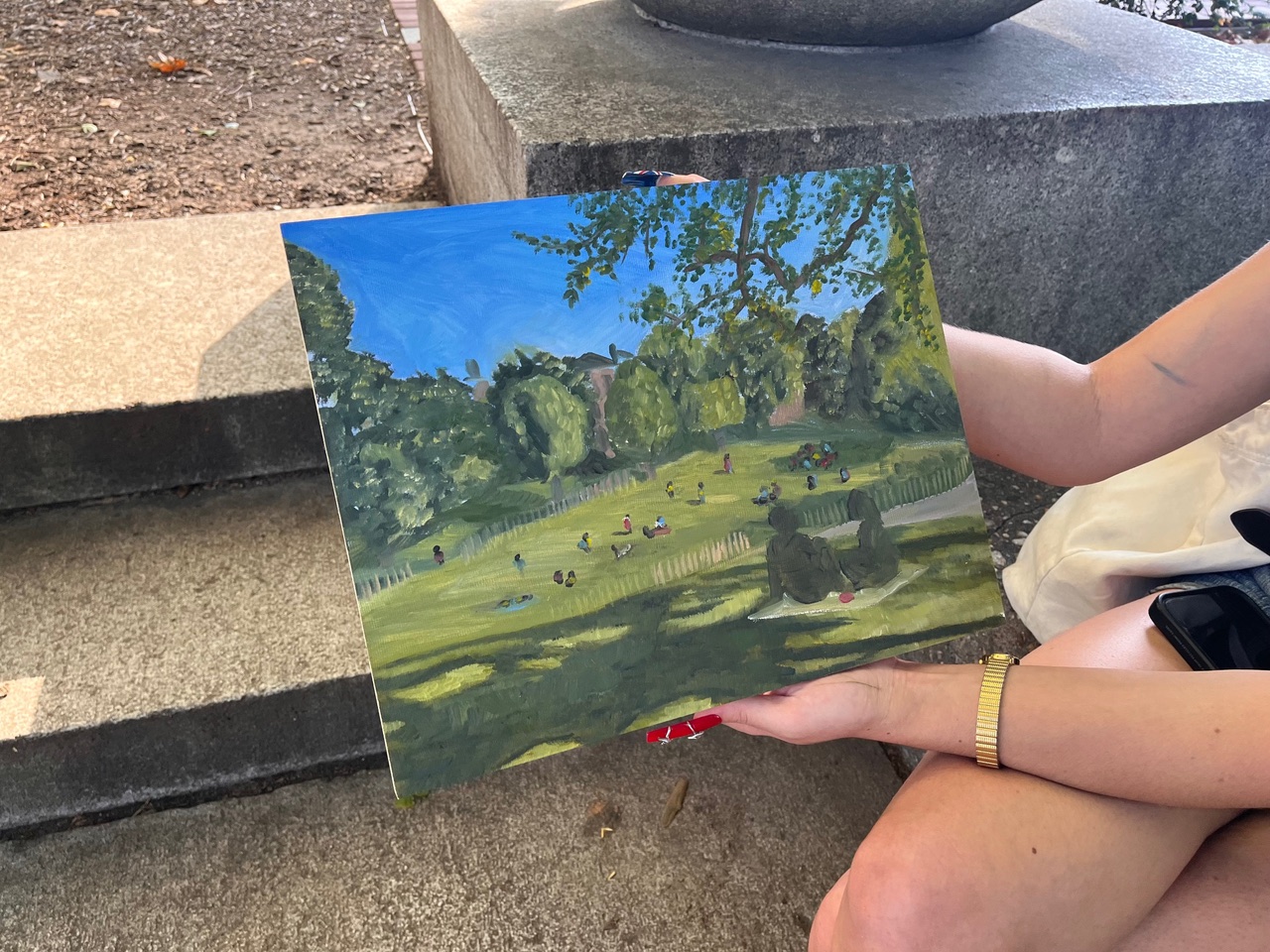 A person sits on the ground next to a large tree, surrounded by fallen leaves and small plants. They are holding a paintbrush and looking at a canvas. The individual is wearing a green jacket and pants, with brown hair styled in a bob. A small art supply bag is beside them on the ground. The scene is set in a natural area with a few trees in the background.