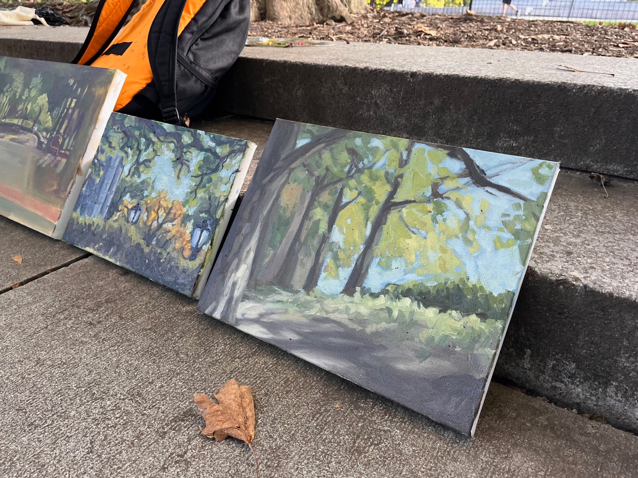 Three paintings are propped against a stone step, featuring various outdoor scenes with trees, foliage, and soft colors. A fallen leaf sits on the ground in front of the canvases. In the background, a person with an orange and black backpack is partially visible.