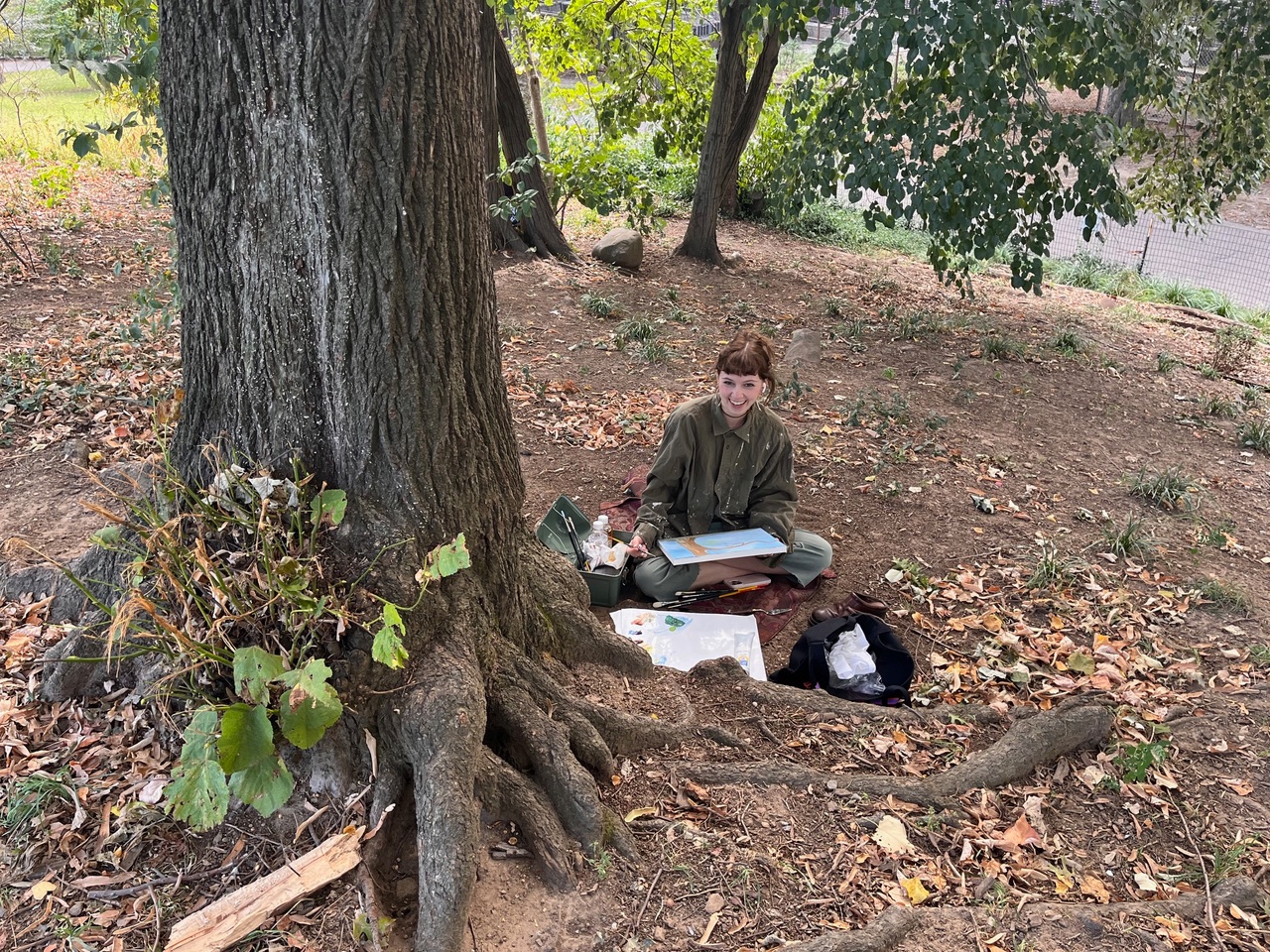 A young person sits on the ground against a large tree, smiling while holding an art piece. They are surrounded by fallen leaves and grass, with a small bag and art supplies nearby. The setting appears to be a peaceful outdoor area.