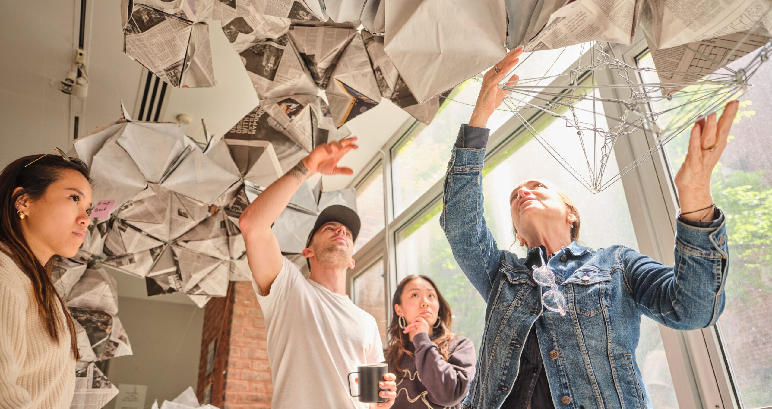 A group of four people are engaged in an activity involving hanging sculptures made from folded newspaper and wire. One person is reaching up to describe the wire structure, while another observes with interest. The setting features large windows allowing natural light to enter, highlighting the creative environment.