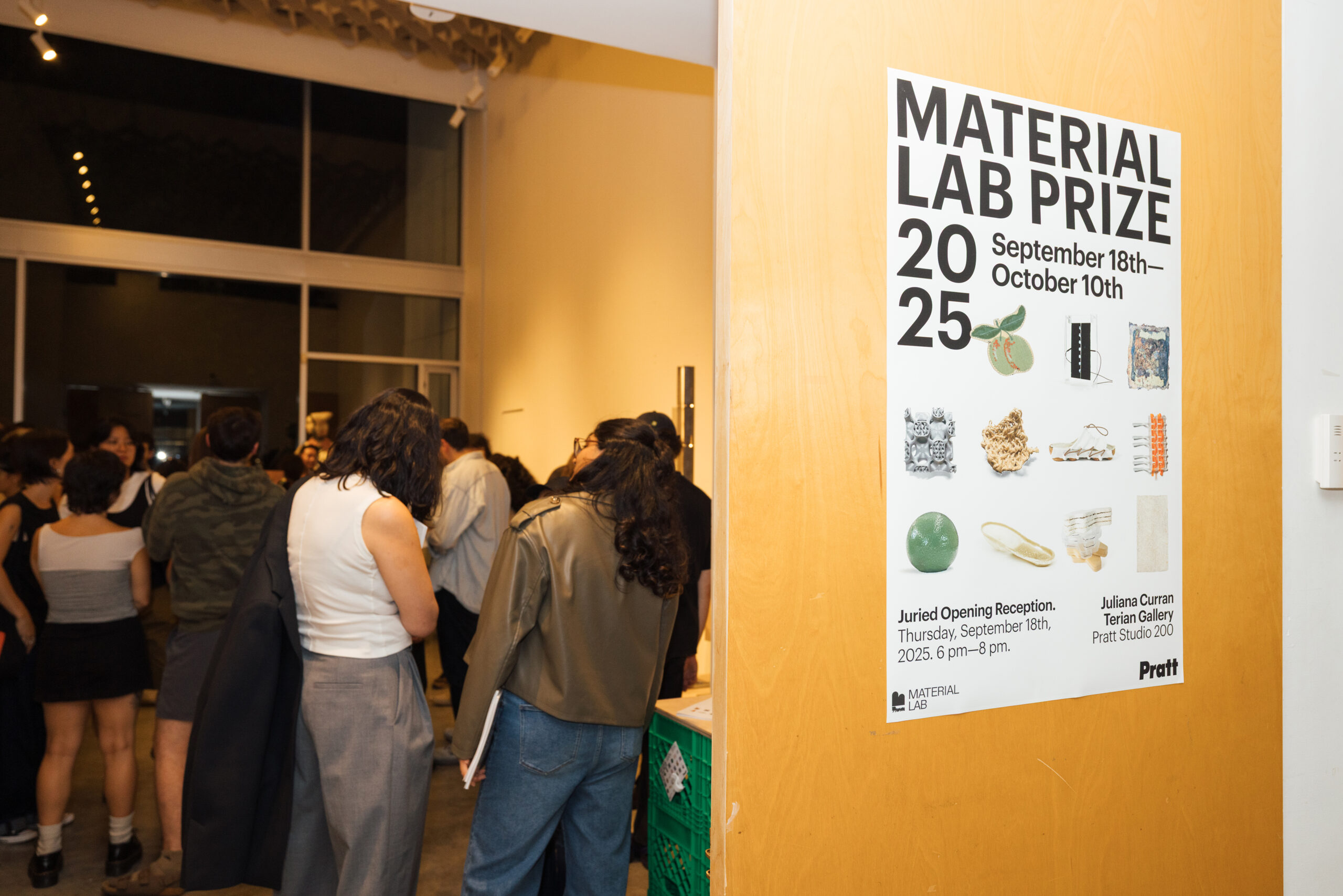 Two young women are interacting with a wooden structure on a table in a brightly lit room. The woman on the left, with curly hair, is looking at the structure, while the woman on the right, wearing a striped shirt, smiles as she places her hand on it. Behind them are shelves and other people engaged in activity.