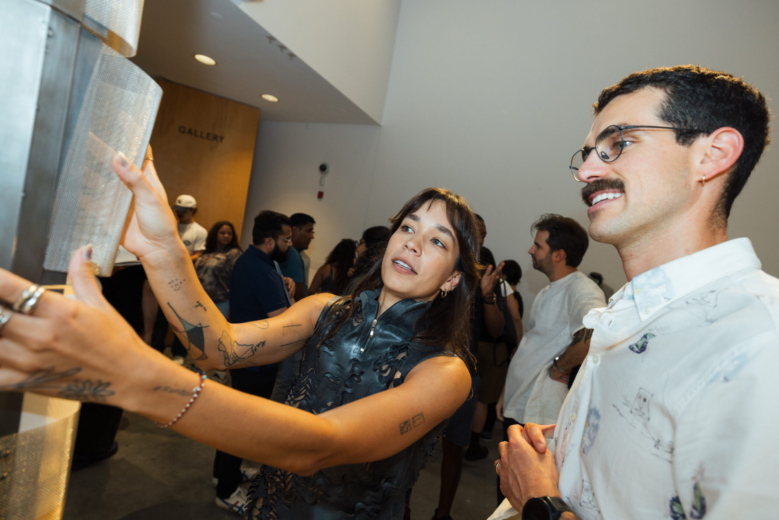 A woman with long hair and tattoos interacts with a metallic display, appearing engaged and animated. She wears a sleeveless, textured outfit. A man with glasses and a mustache stands beside her, smiling and observing the interaction. In the background, several other people can be seen, contributing to a lively gallery atmosphere. The gallery sign is partially visible.