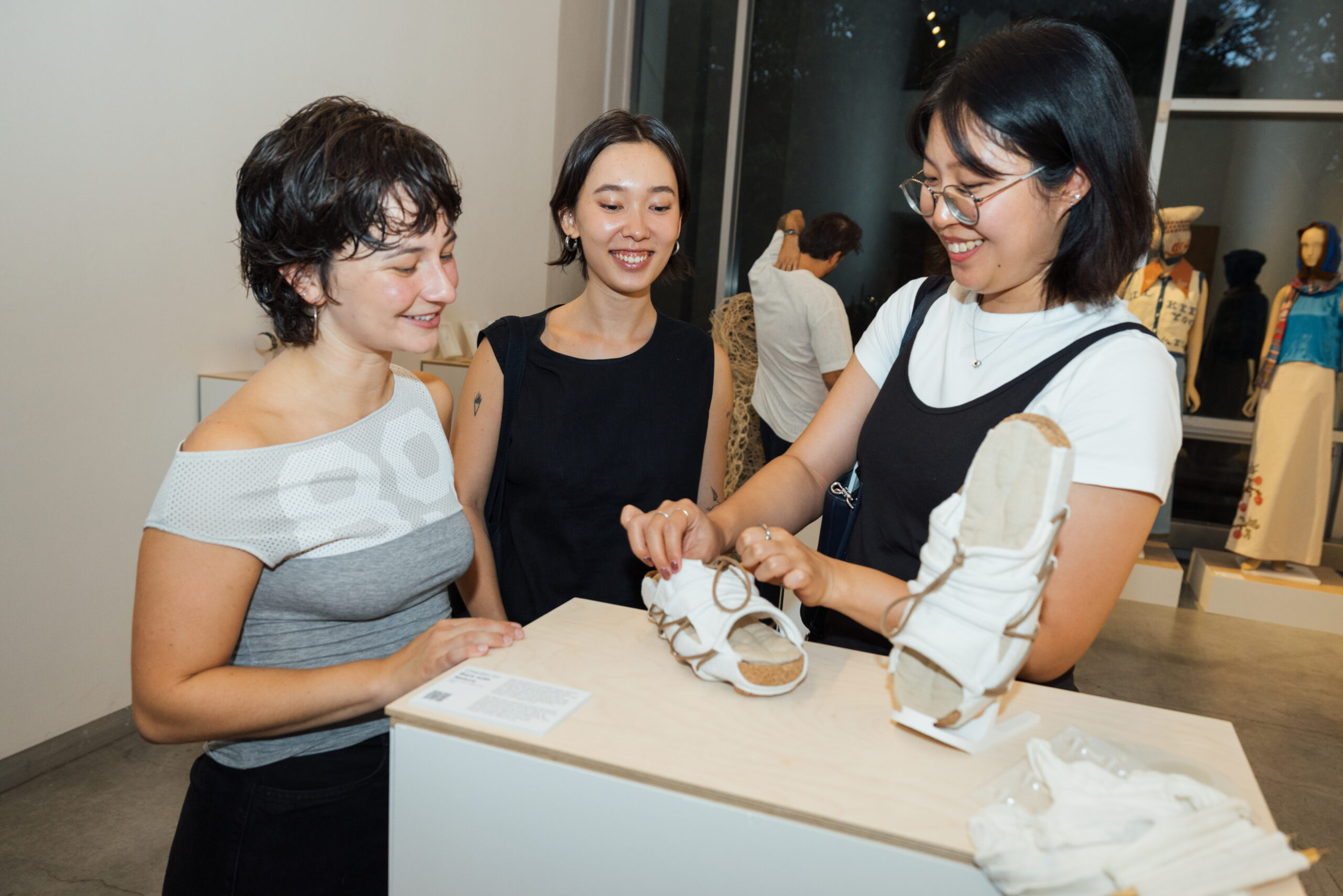 Three women are engaged in conversation at an art exhibition. One woman, dressed in a gray off-shoulder top, smiles while leaning on a display table. The second woman, wearing a black sleeveless dress, looks on with a smile. The third woman, in a black and white outfit, examines a pair of unique white sandals on the table. In the background, additional artworks can be seen, including clothing displays.