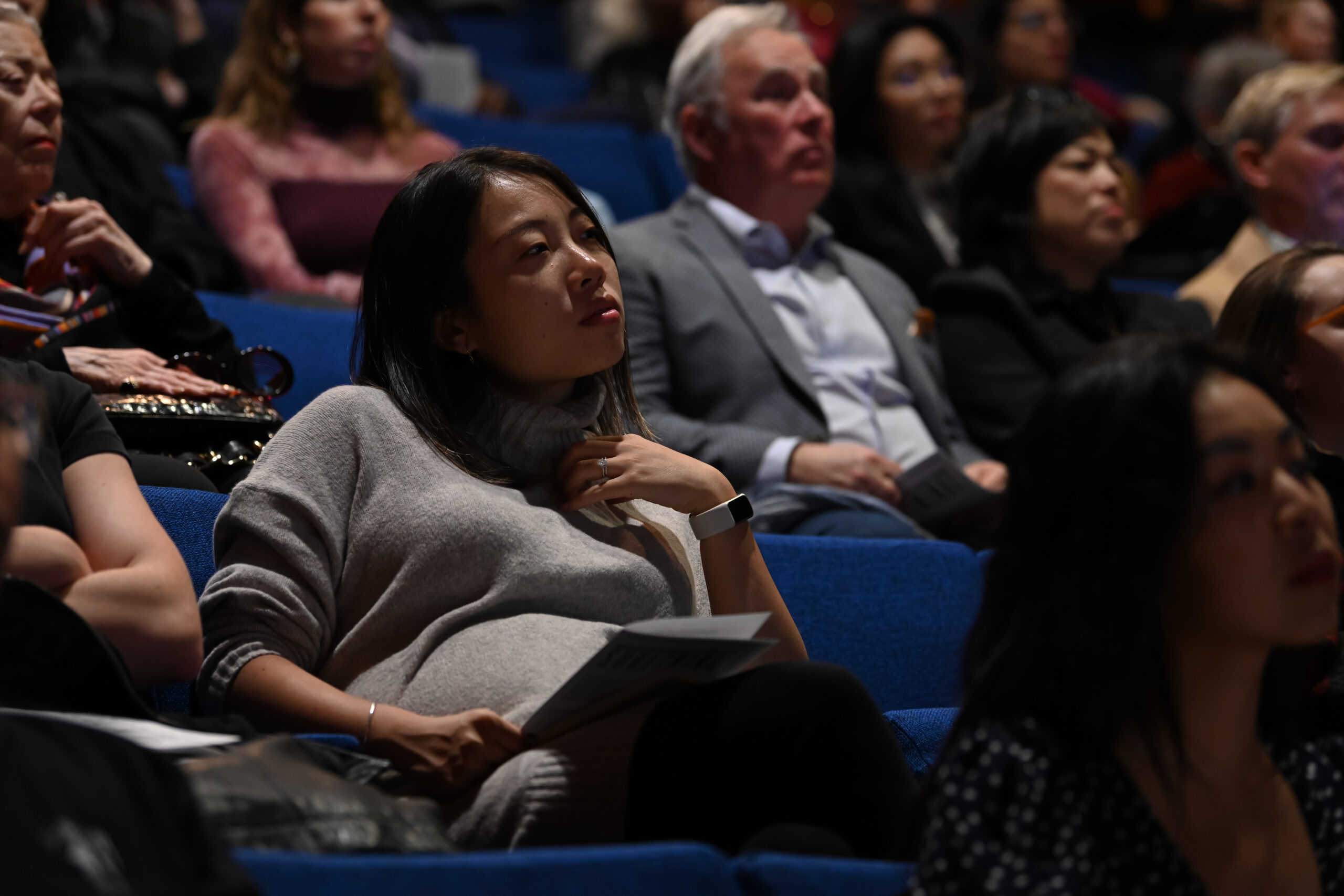 A crowd of people is seated in a theater, with blue seating in the background. A woman in a gray sweater is seated in the foreground, looking thoughtful while holding a program. Other audience members are visible behind her, with varying expressions, engaged in the presentation.