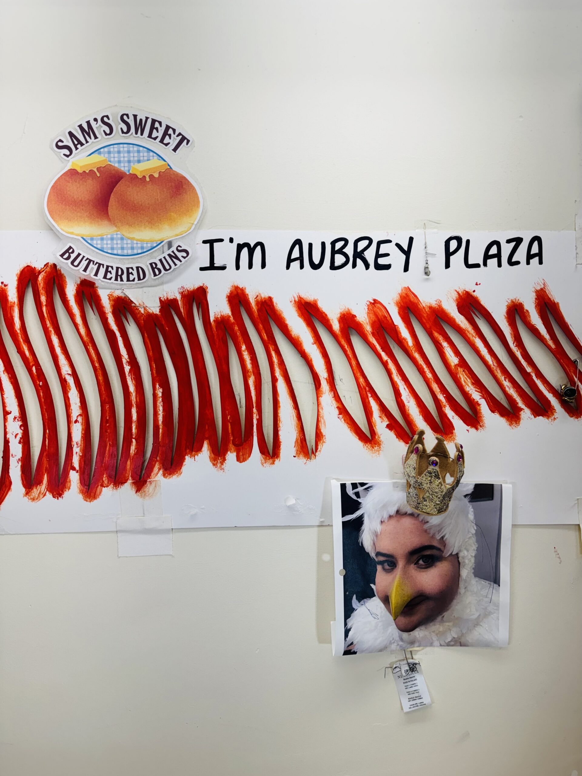 A wall hung with various pieces of ephemera, including a sign reading Sam's Hot Buttered Buns, a striped stencil cut from a cue card reading "I'm Aubrey Plaza," and a close-up portrait of an actor wearing a chicken costume, topped with a tiny gold crown