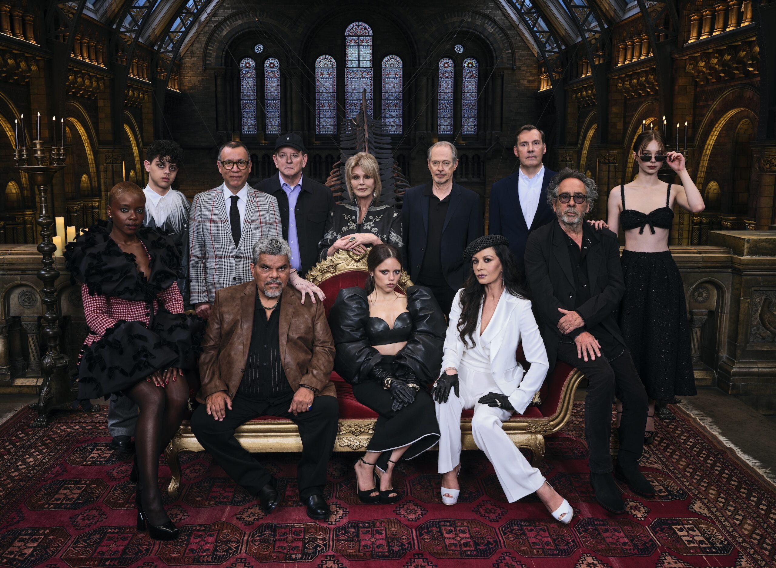 Promotional photo of an ensemble cast of actors dressed in sharp, mainly black-and-white attire assembled at the Natural History Museum in London