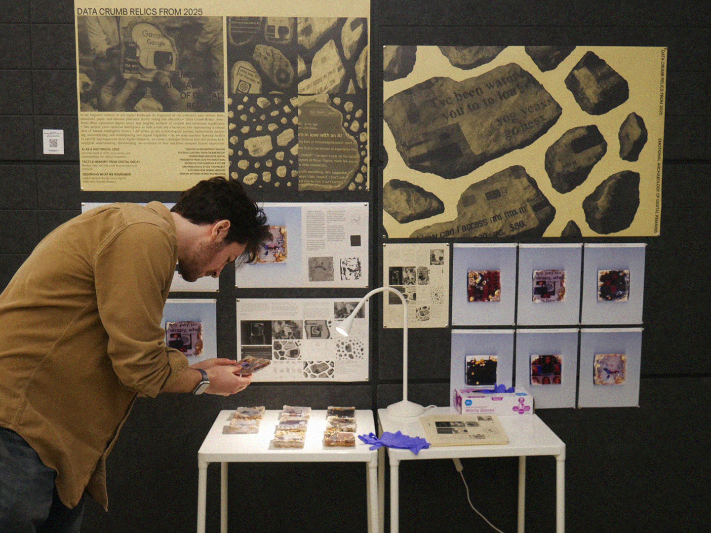 A person in a brown shirt examines items on a table under a light, focused on collectibles arranged in a row. Behind them is a wall displaying several posters with text and images related to "Data Crumb Relics from 2025." The setup includes a small stack of gloves and a document on the table.